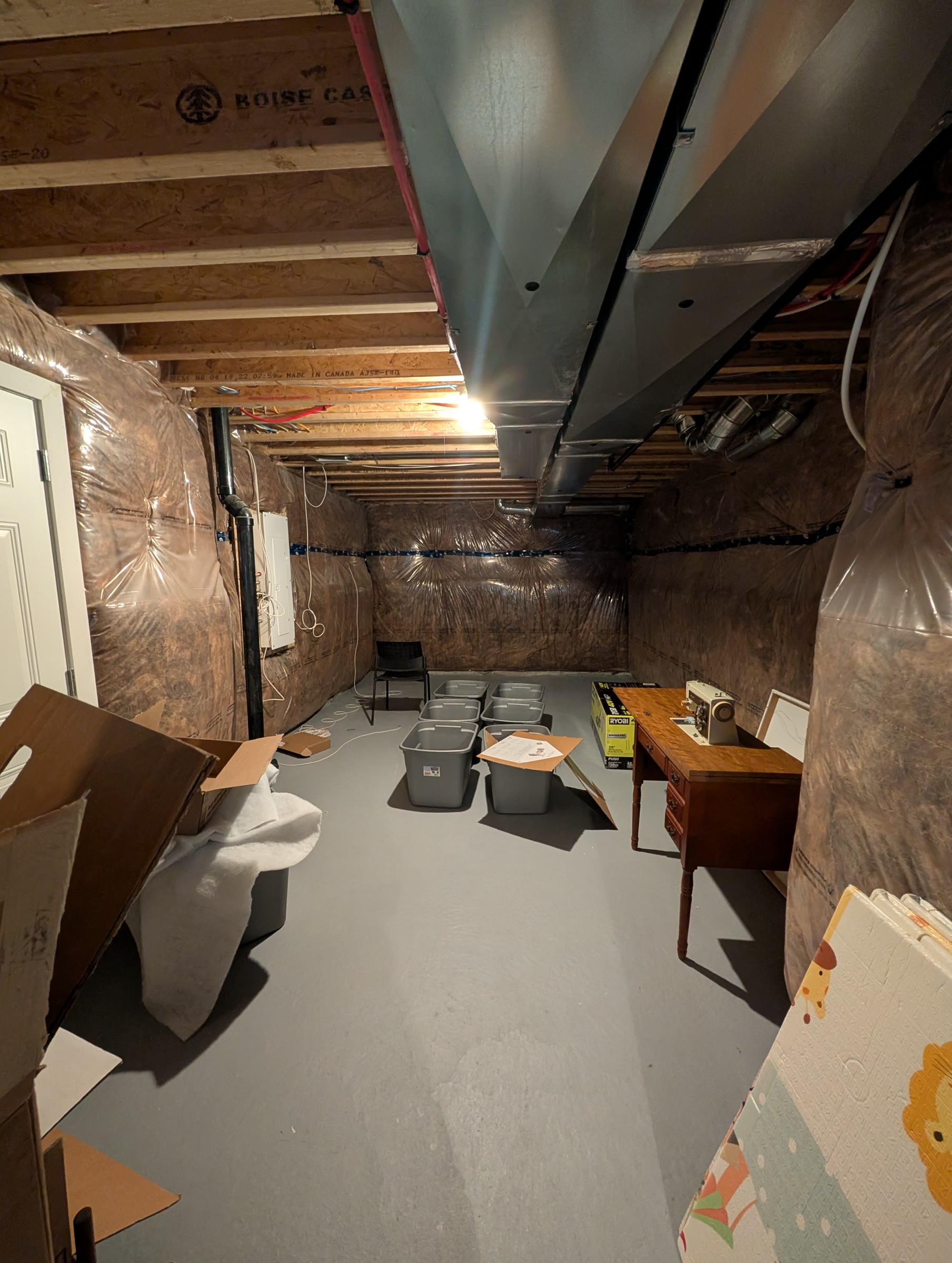Empty unfinished basement with exposed joists, wrapped insulation, storage bins, and a sewing table before the makeover