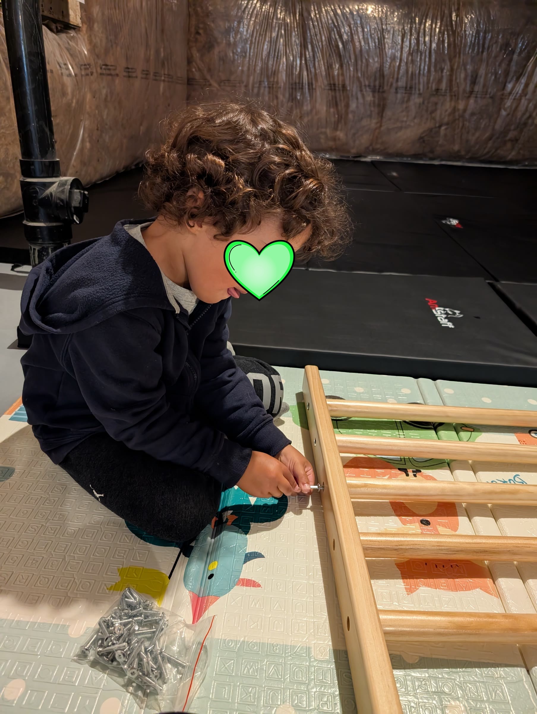 A toddler sitting on a play mat helping screw a wooden ladder frame together, with a bag of screws nearby