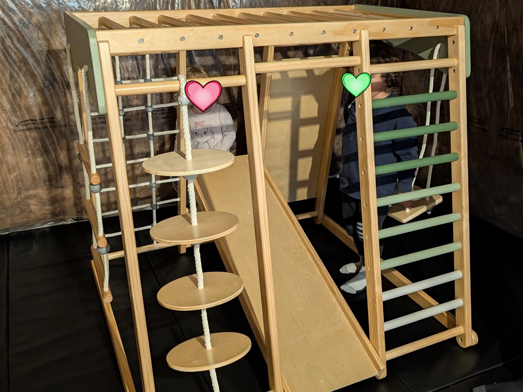Wooden jungle gym with a slide, climbing net, rope steps, and ladder on black gym mats in a basement play area