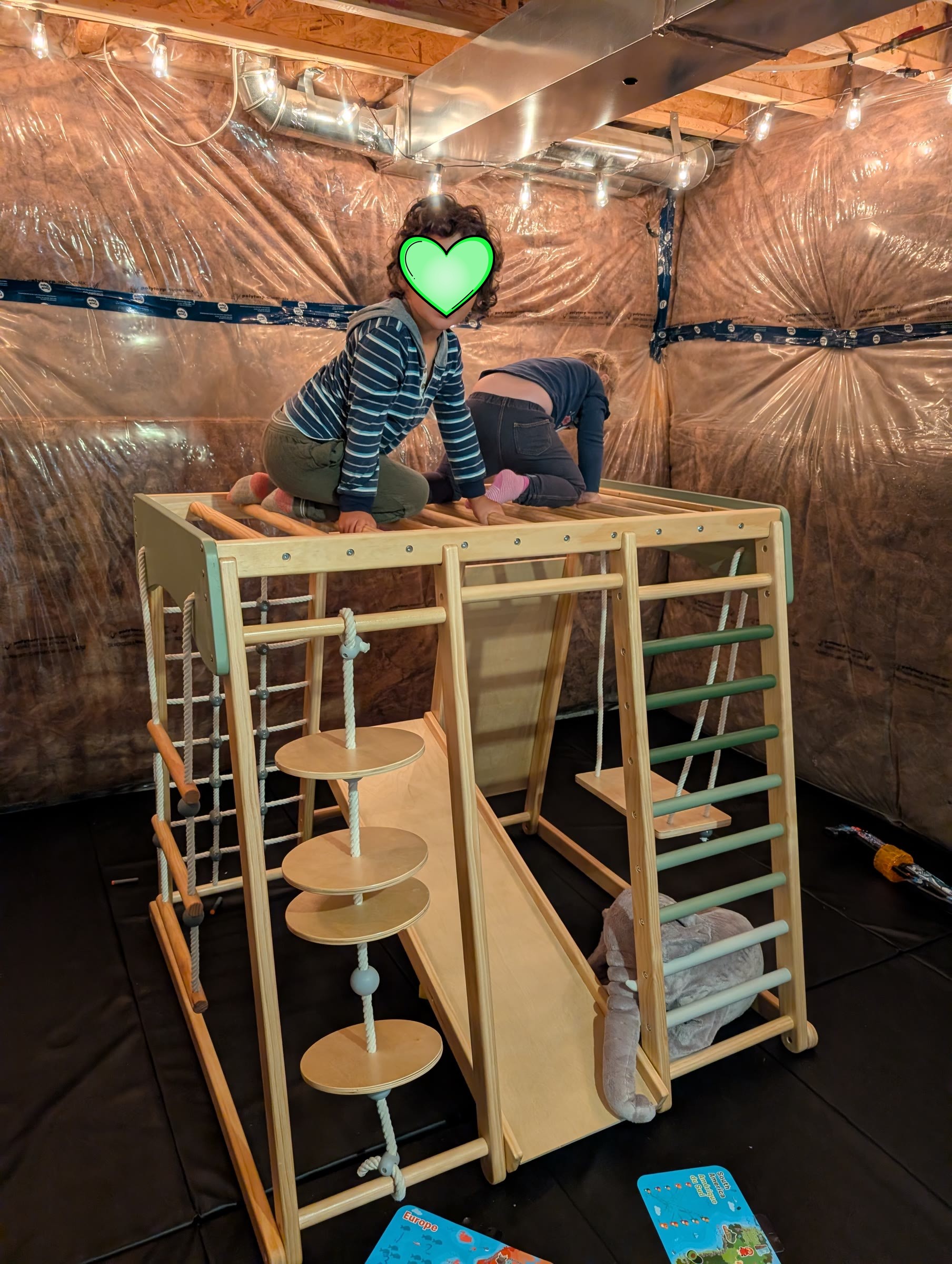 Two kids climbing on a wooden jungle gym on black gym mats in a basement lit with Edison string lights