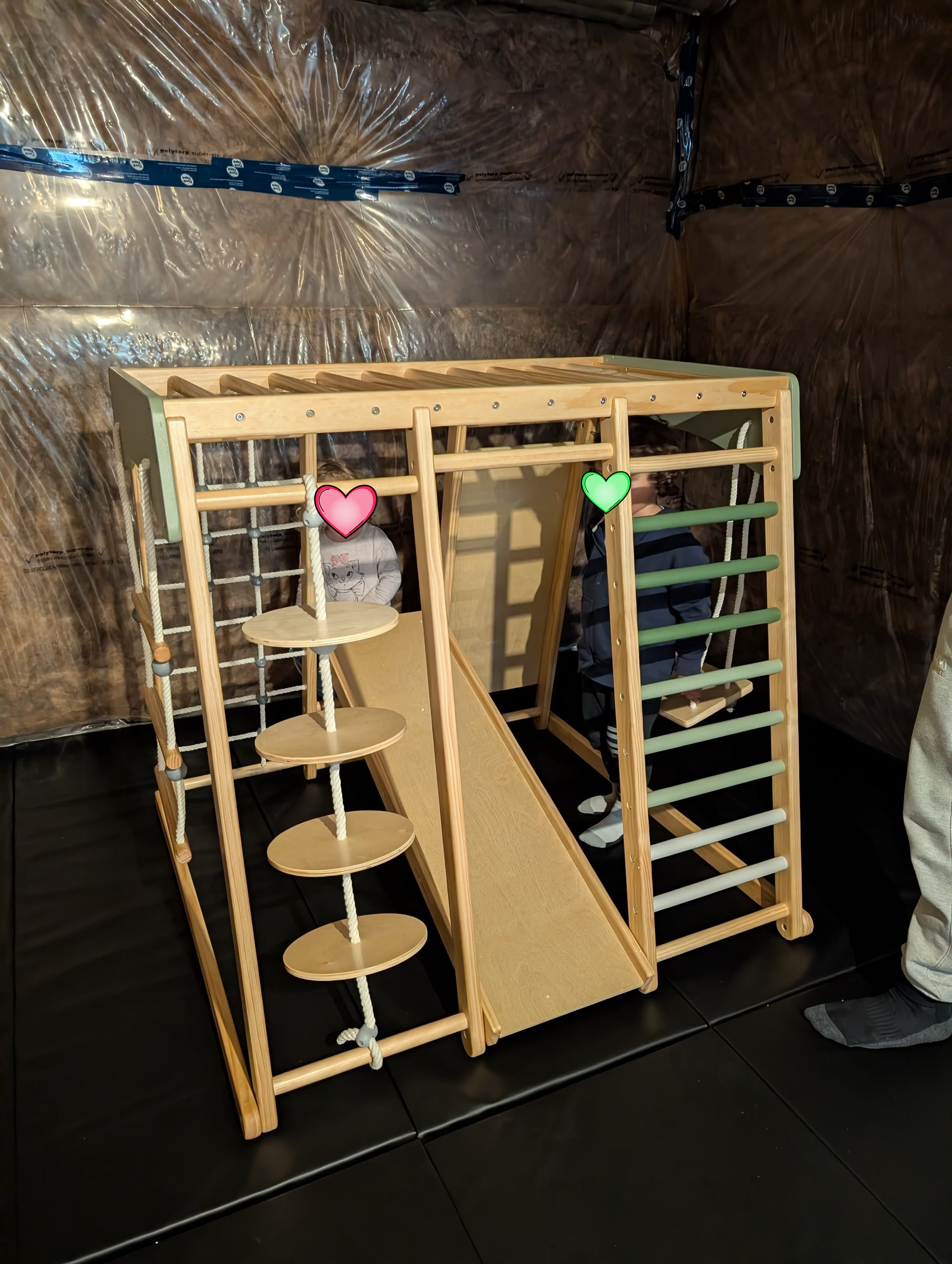 Fully assembled wooden jungle gym with a slide, climbing net, rope steps, and ladder on black gym mats, with two kids standing inside it