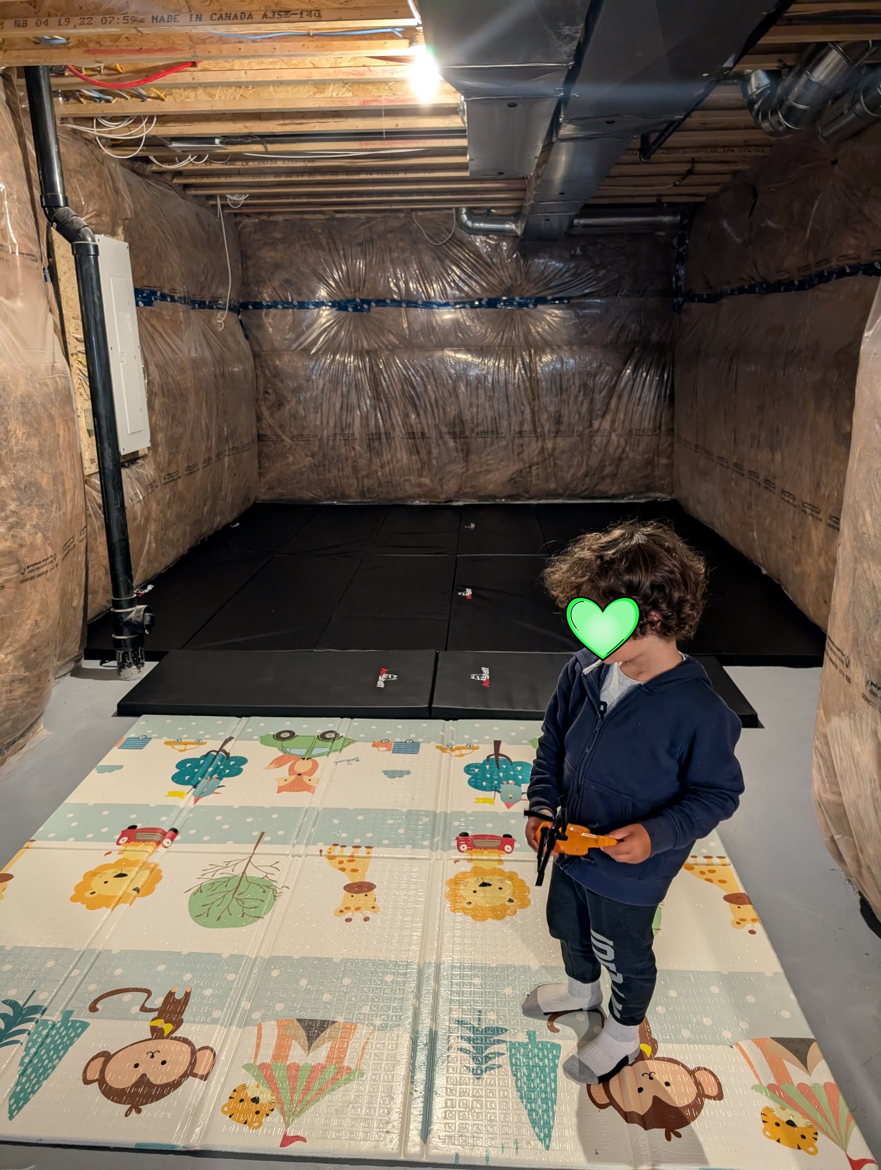 Black gym mats laid across the basement floor with a colorful foam play mat in front and a toddler standing on it