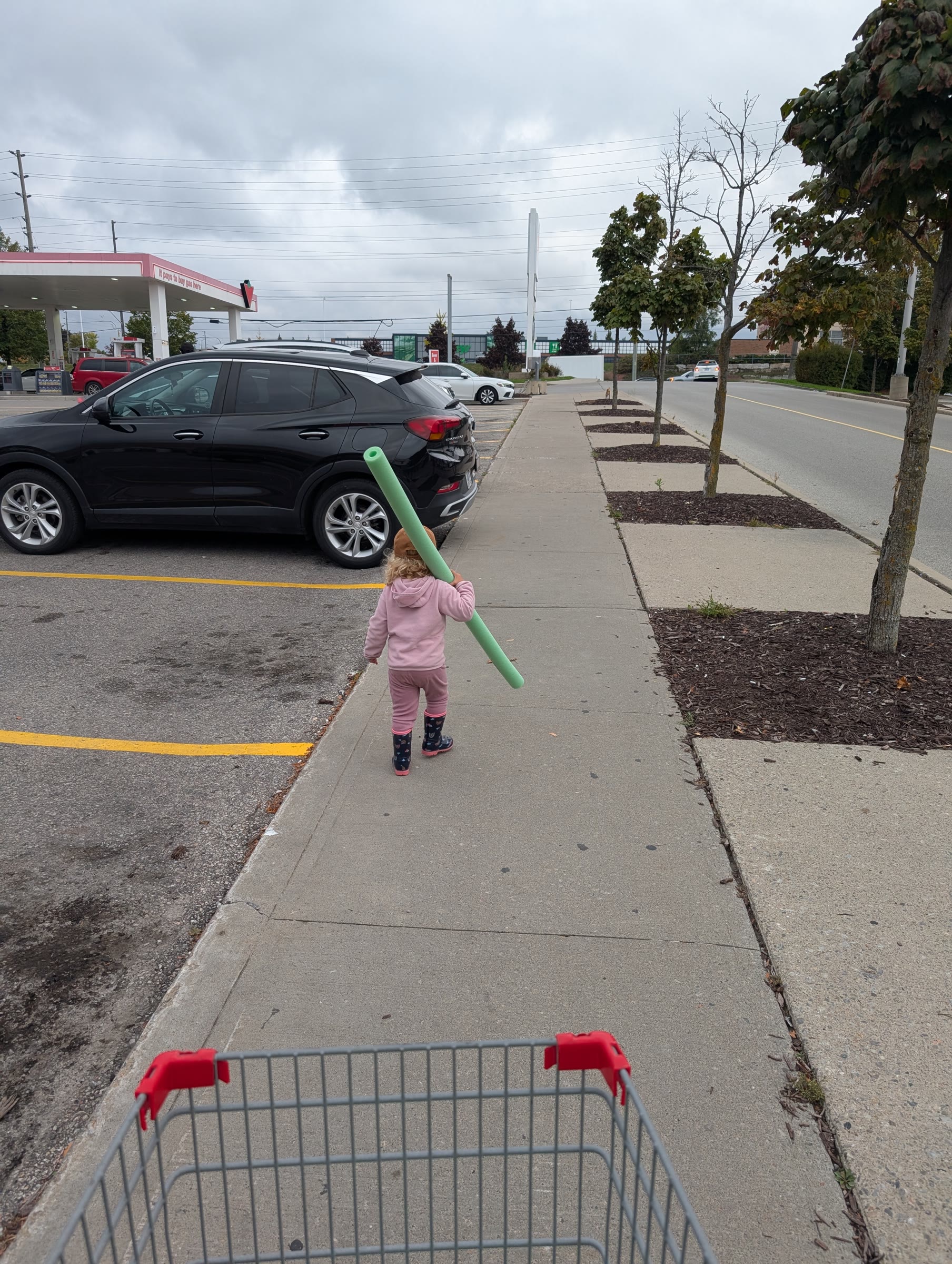 A toddler in a pink hoodie and rain boots carrying a green pool noodle over one shoulder along a sidewalk