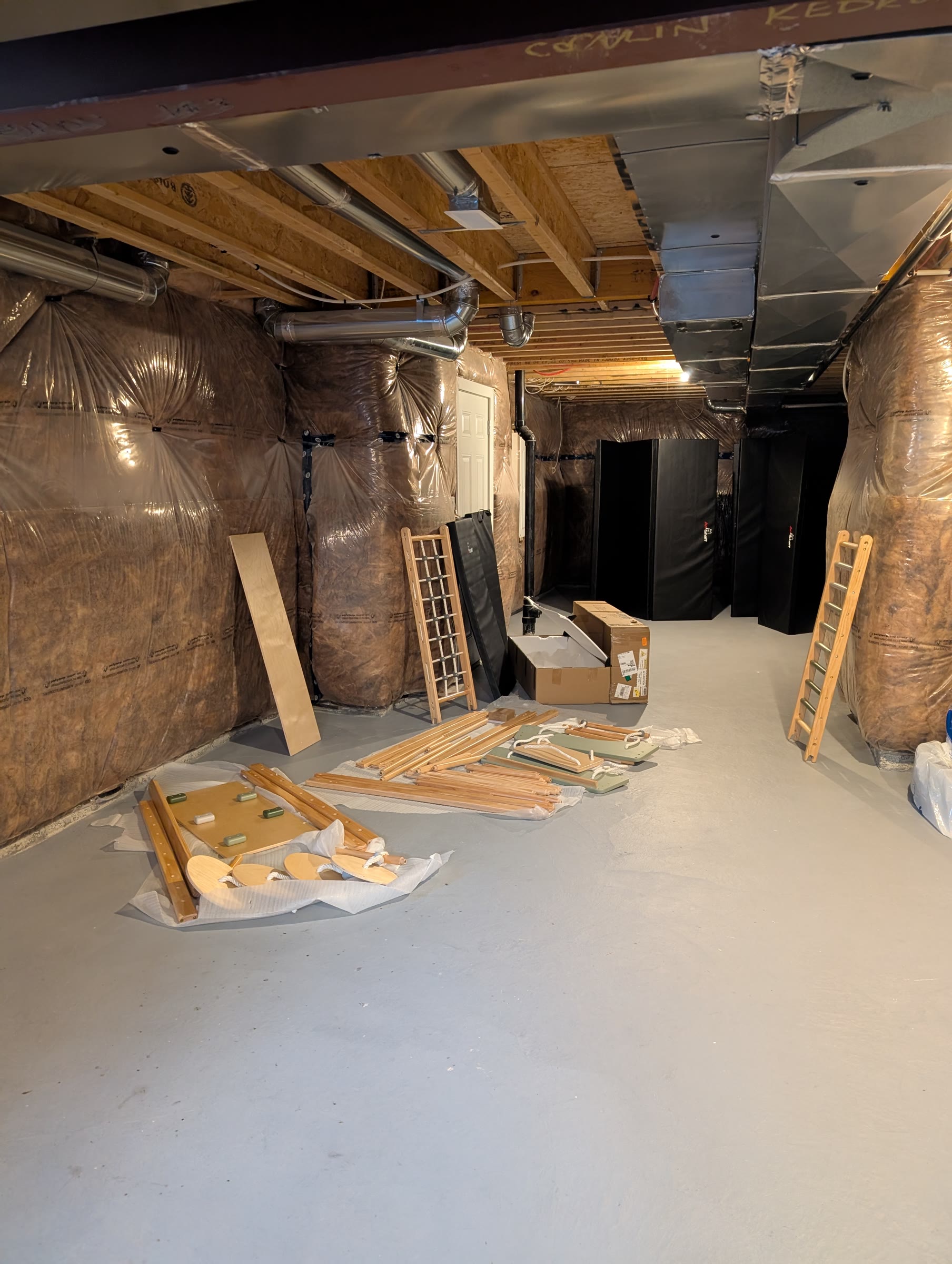 Wooden jungle gym pieces, ladders, and swing boards laid out on the basement floor next to the box