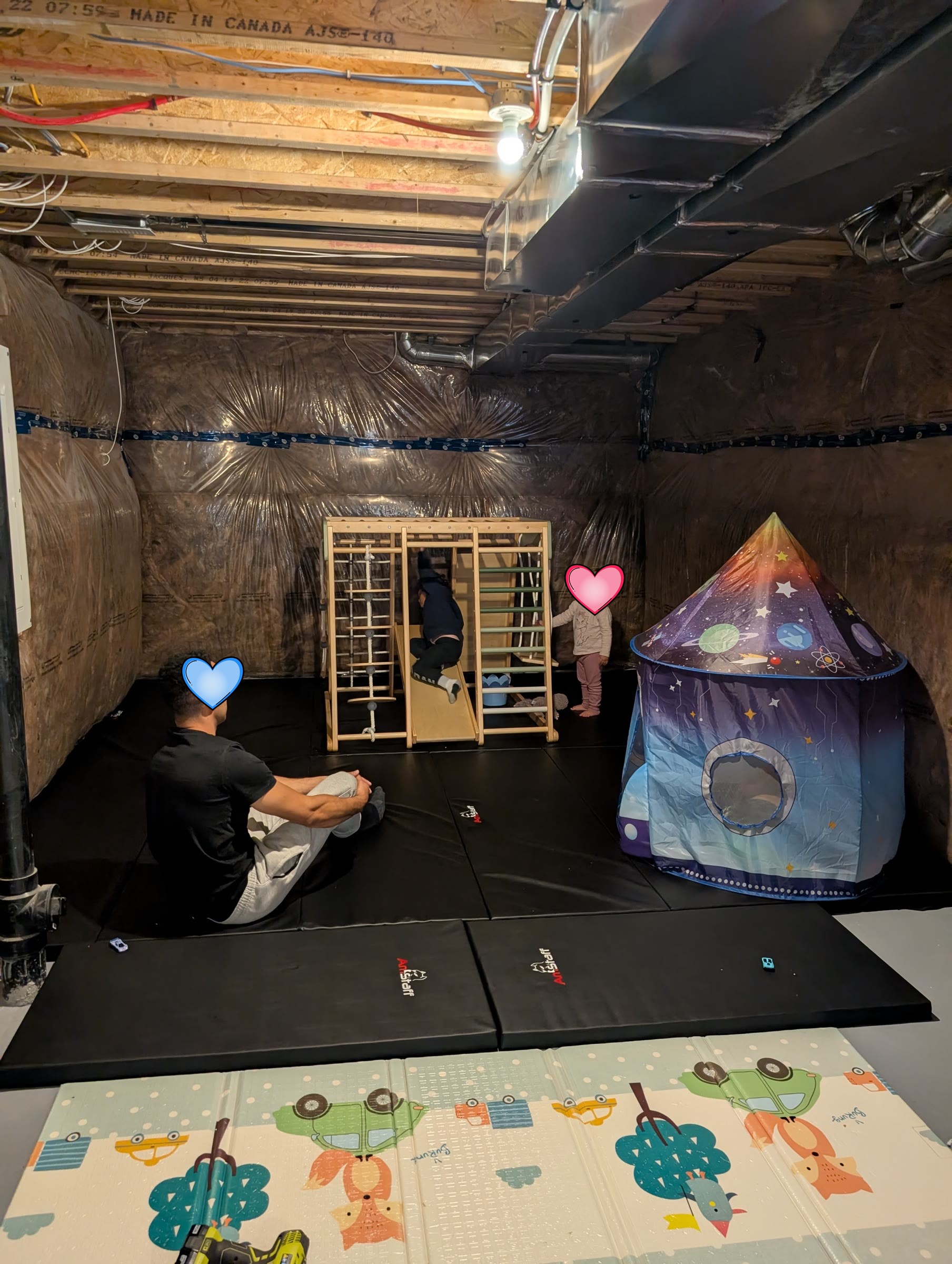 Wide view of the basement play area with the jungle gym, a space-themed play tent, black gym mats, and a parent sitting on the floor