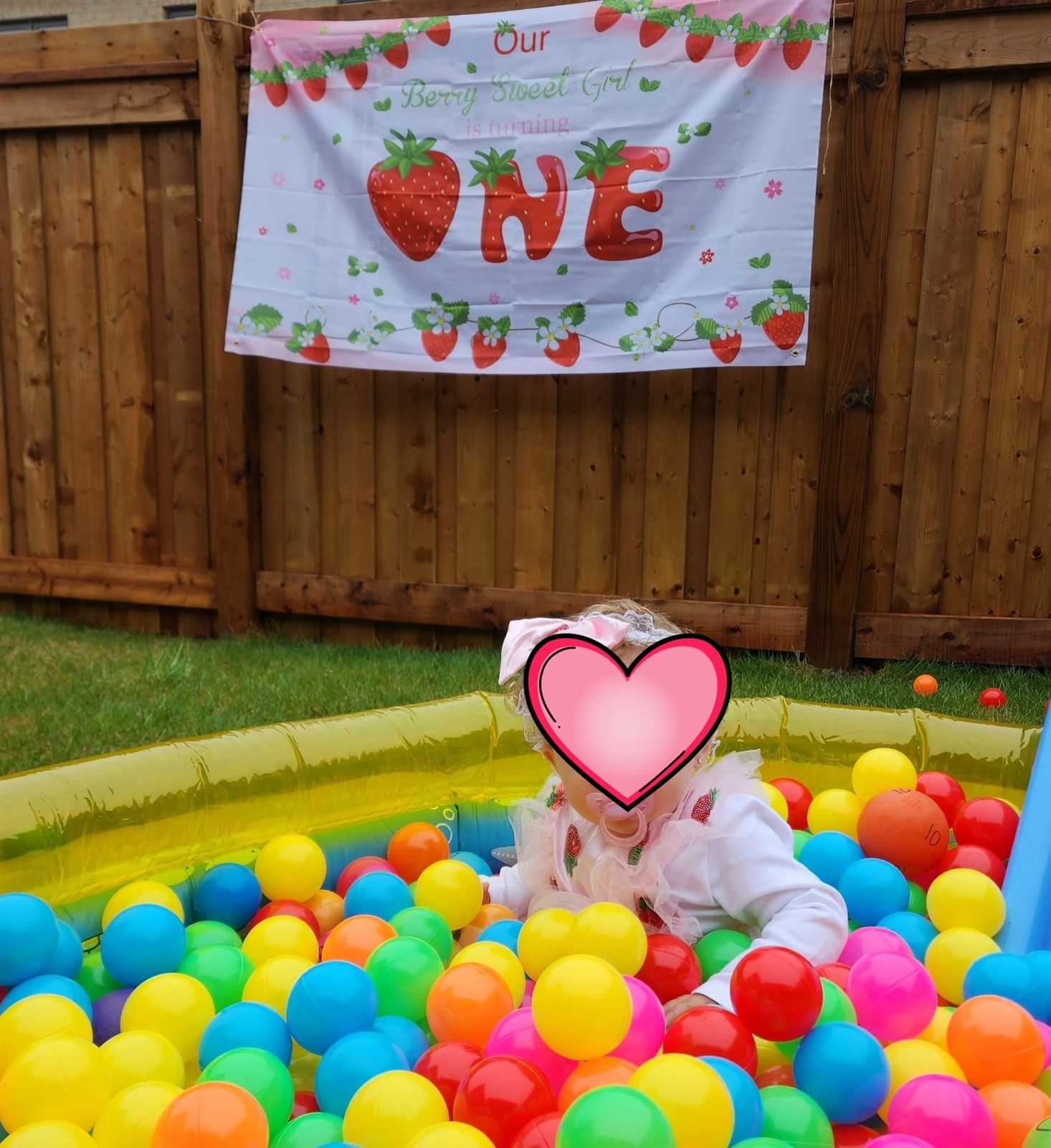 Birthday girl in a ball pit under a strawberry ONE banner hung on the backyard fence