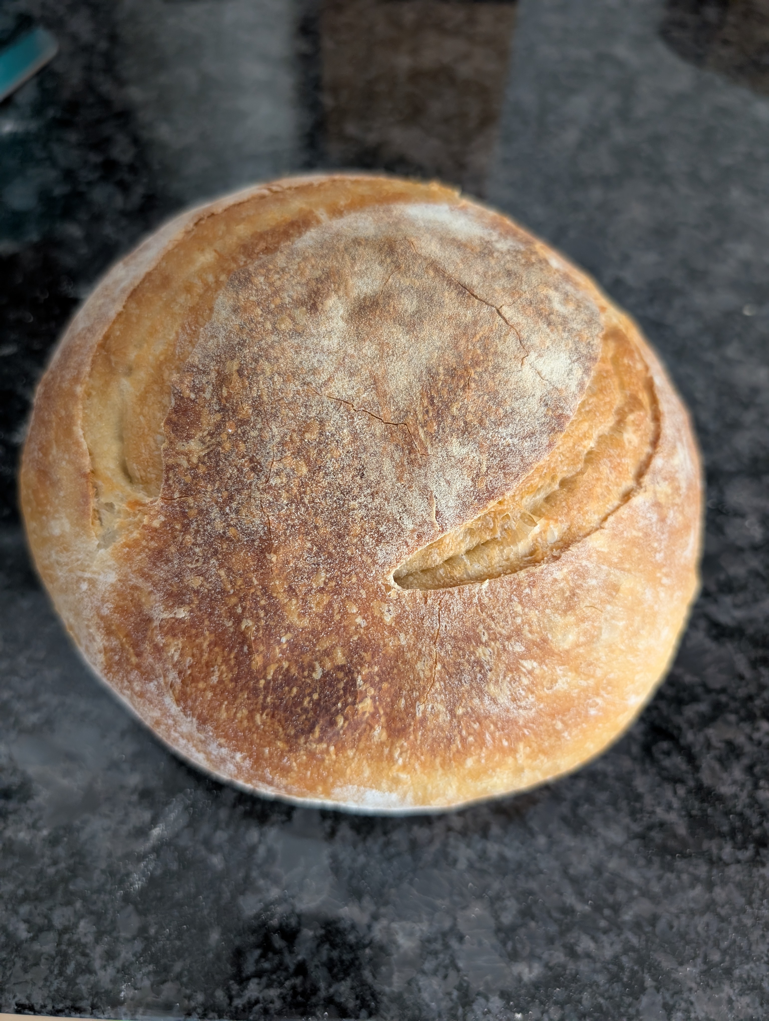 Finished round sourdough loaf with a golden, flour-dusted crust and a scored ear, sitting on a dark countertop