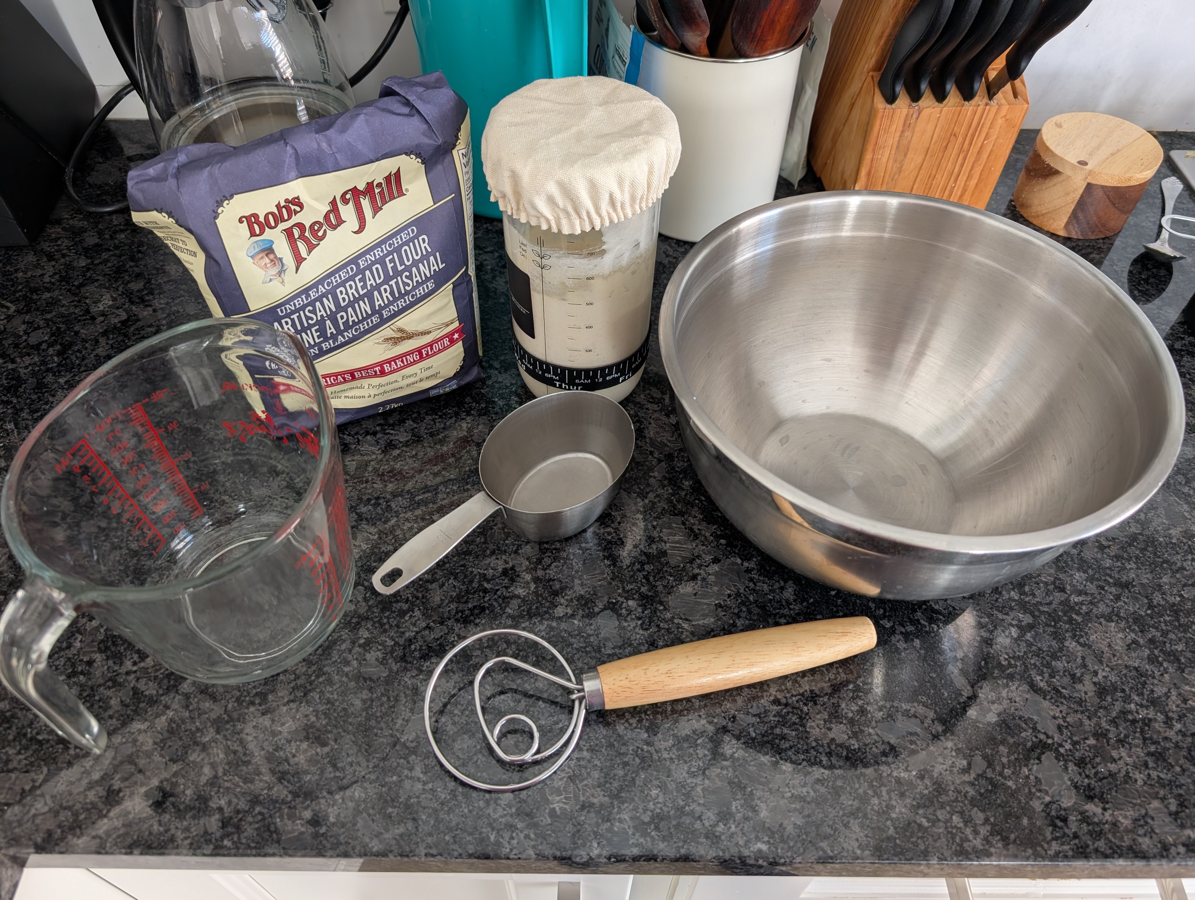 Bread flour, fed starter, measuring cups, a mixing bowl, and a dough whisk laid out on the counter