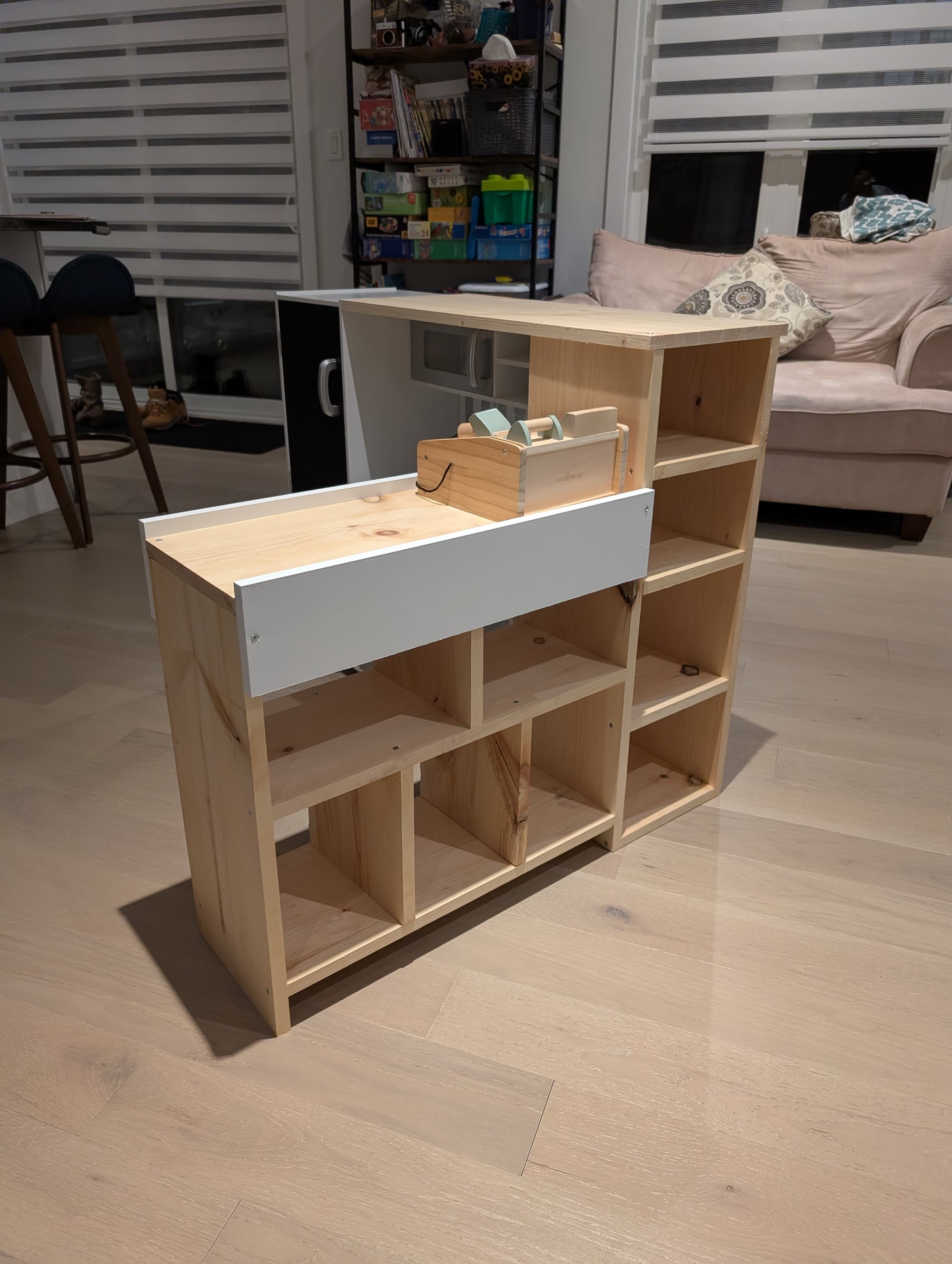 Assembled unfinished pine checkout counter with a white front panel and the wooden cash register sitting on top, next to the play kitchen