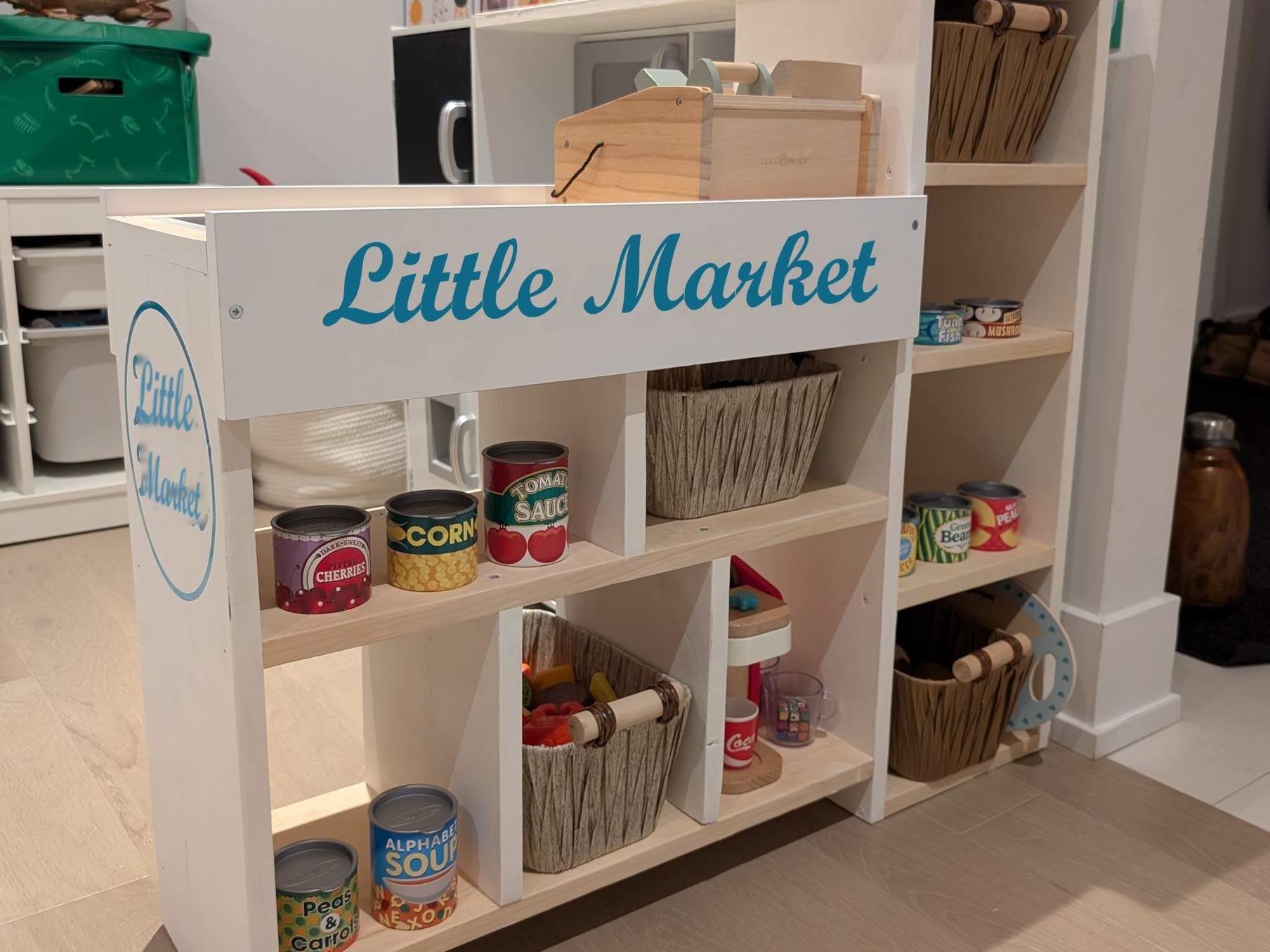 Finished white DIY toddler checkout counter with a Little Market sign, baskets, and play cans, attached to a play kitchen