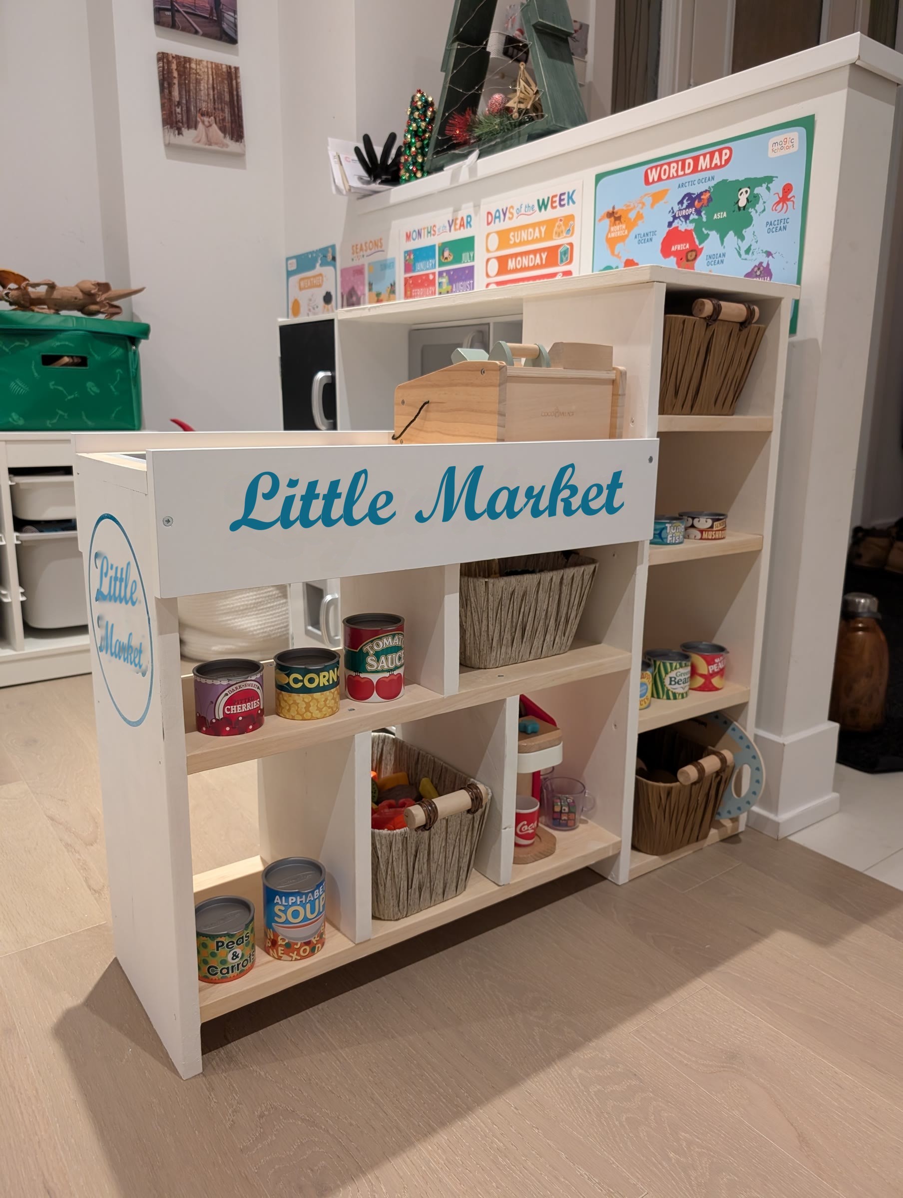 Finished white DIY toddler checkout counter with a Little Market sign, baskets, play cans, and a wooden cash register, attached to a play kitchen