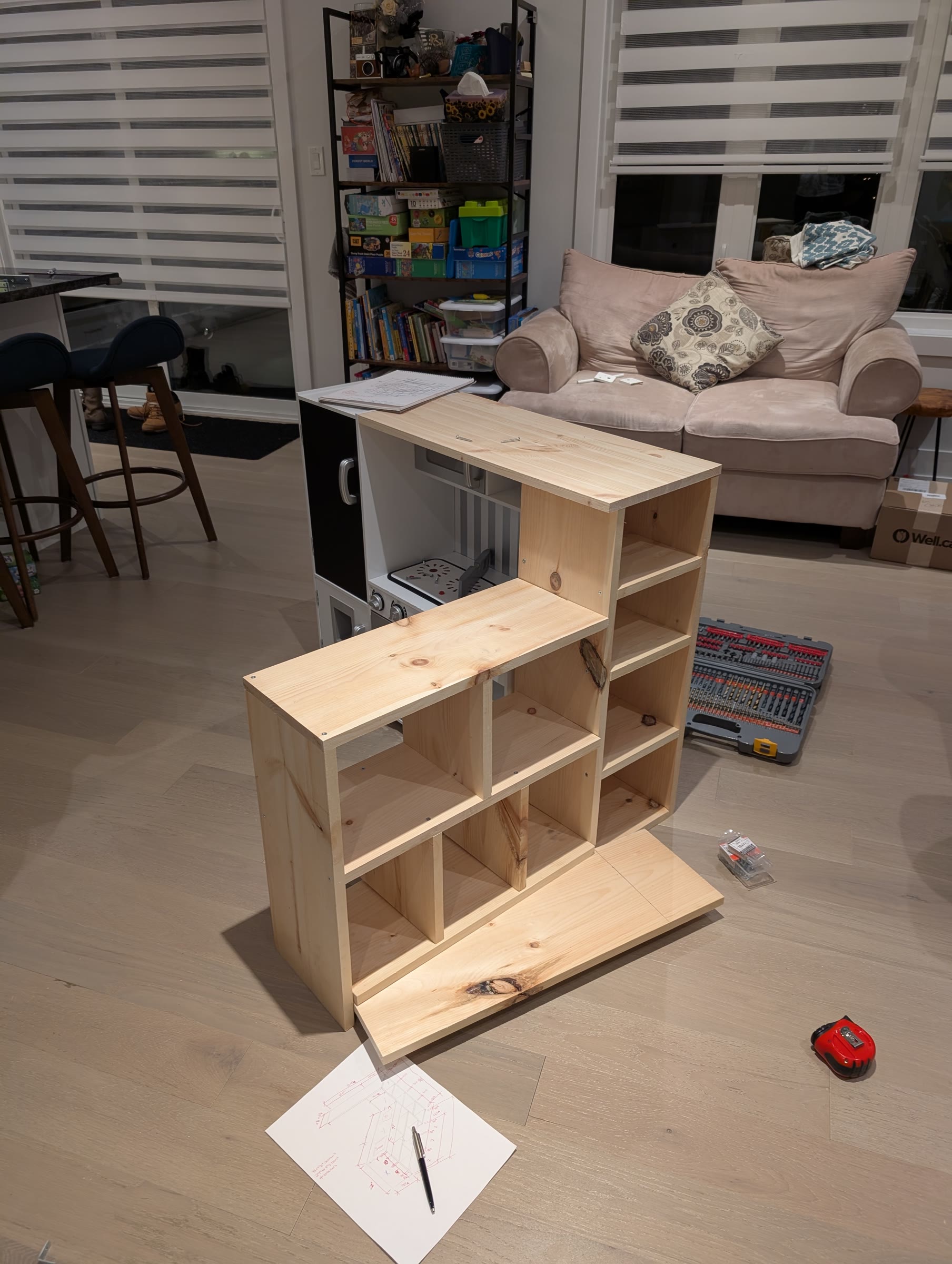 Unfinished pine checkout counter frame assembled next to the play kitchen, with the sketch on top of the kitchen and tools on the floor