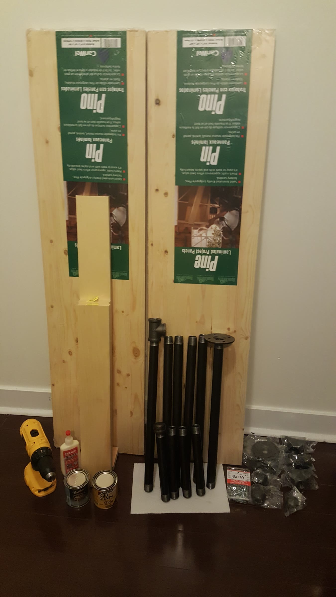 Materials laid out against a wall: two pine laminated project panels, a 1x4 board, black iron pipe and fittings, wood glue, stain, polyurethane, a drill, and screws