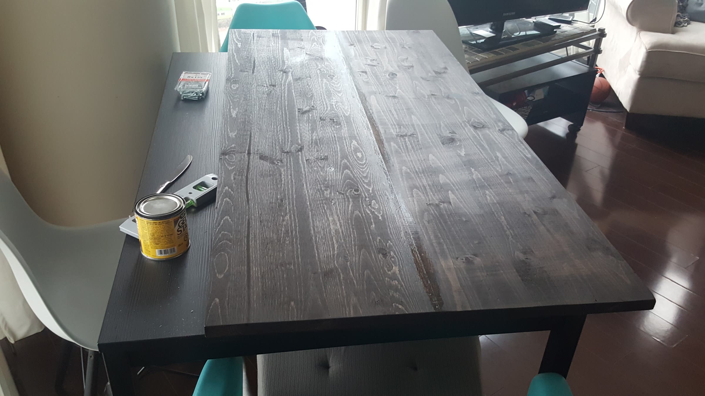 Two stained pine planks laid side by side on a table next to a can of gel stain, a level, and a box of screws