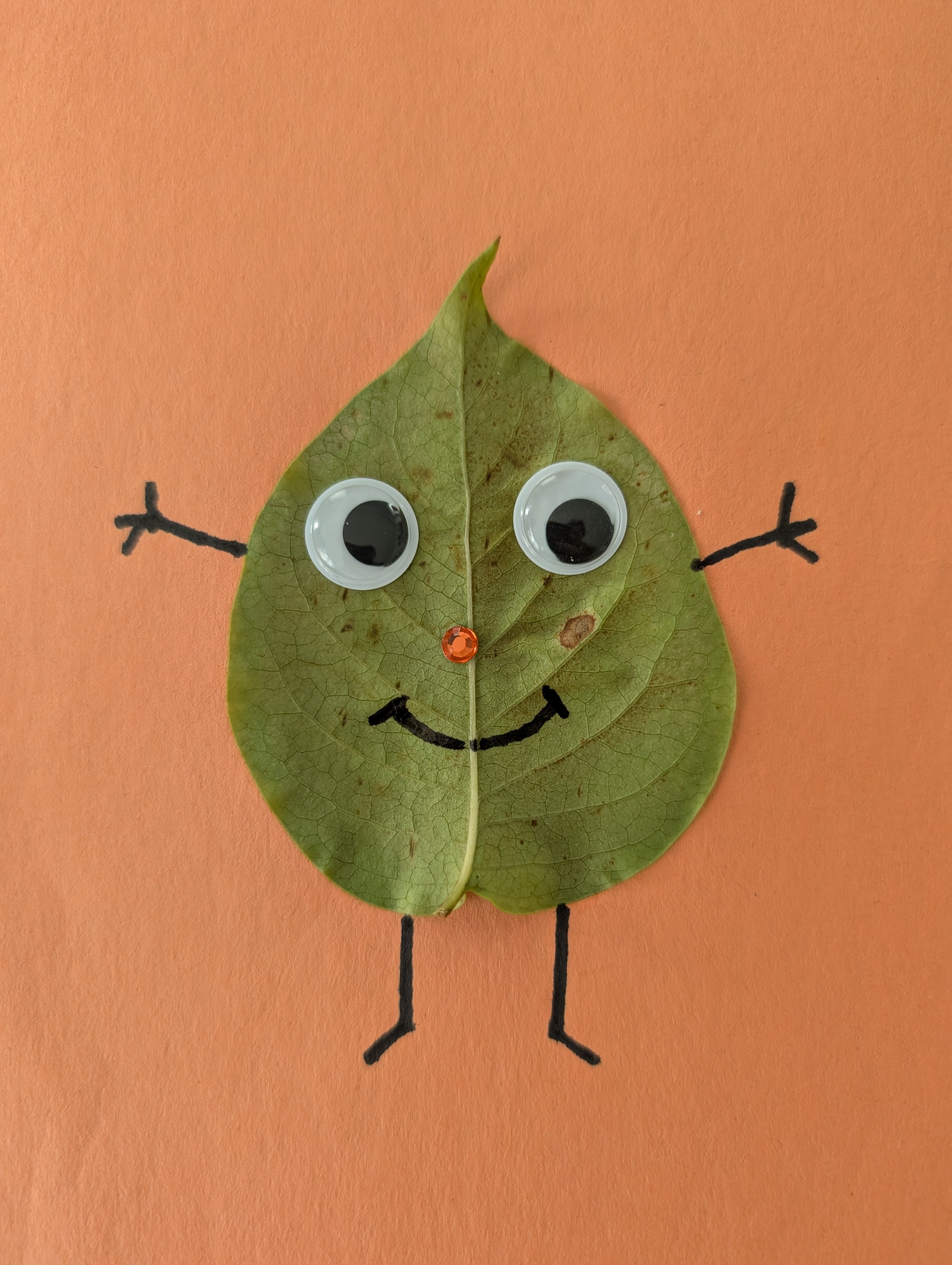 A smiling leaf person with googly eyes, a rhinestone nose, and marker-drawn arms and legs on orange craft paper