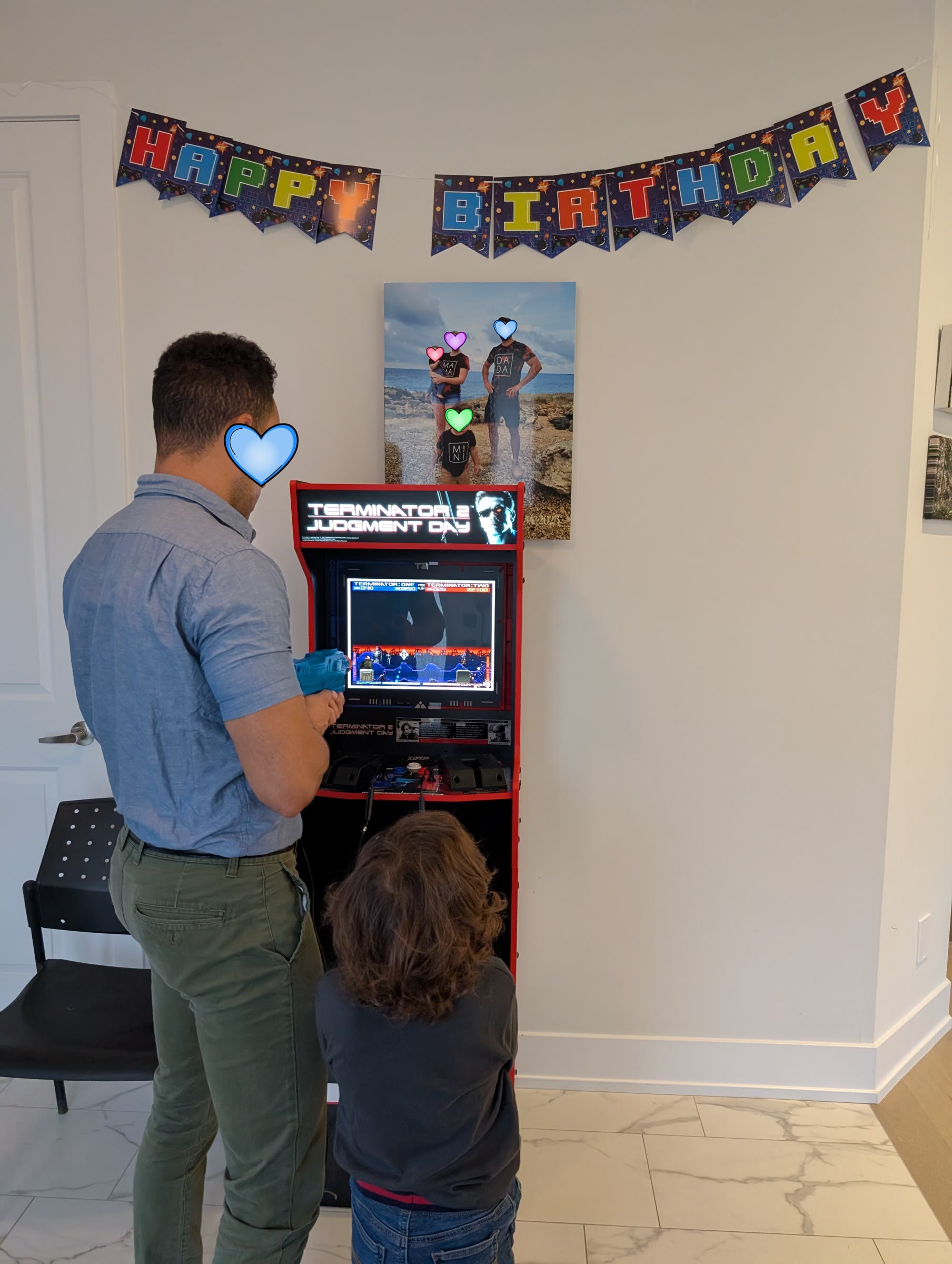 Adult and child playing the Terminator 2 arcade machine under a Happy Birthday video game banner, with coloured hearts covering faces