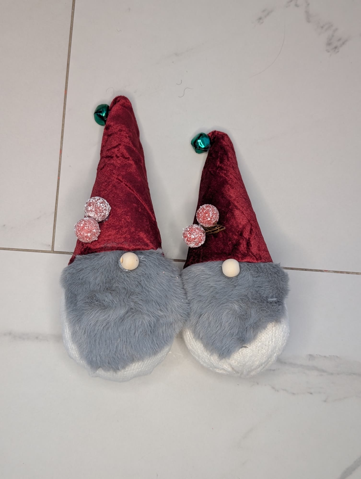 The two finished sock gnomes on a white tile floor, with grey faux fur beards, red velvet hats and wood bead noses