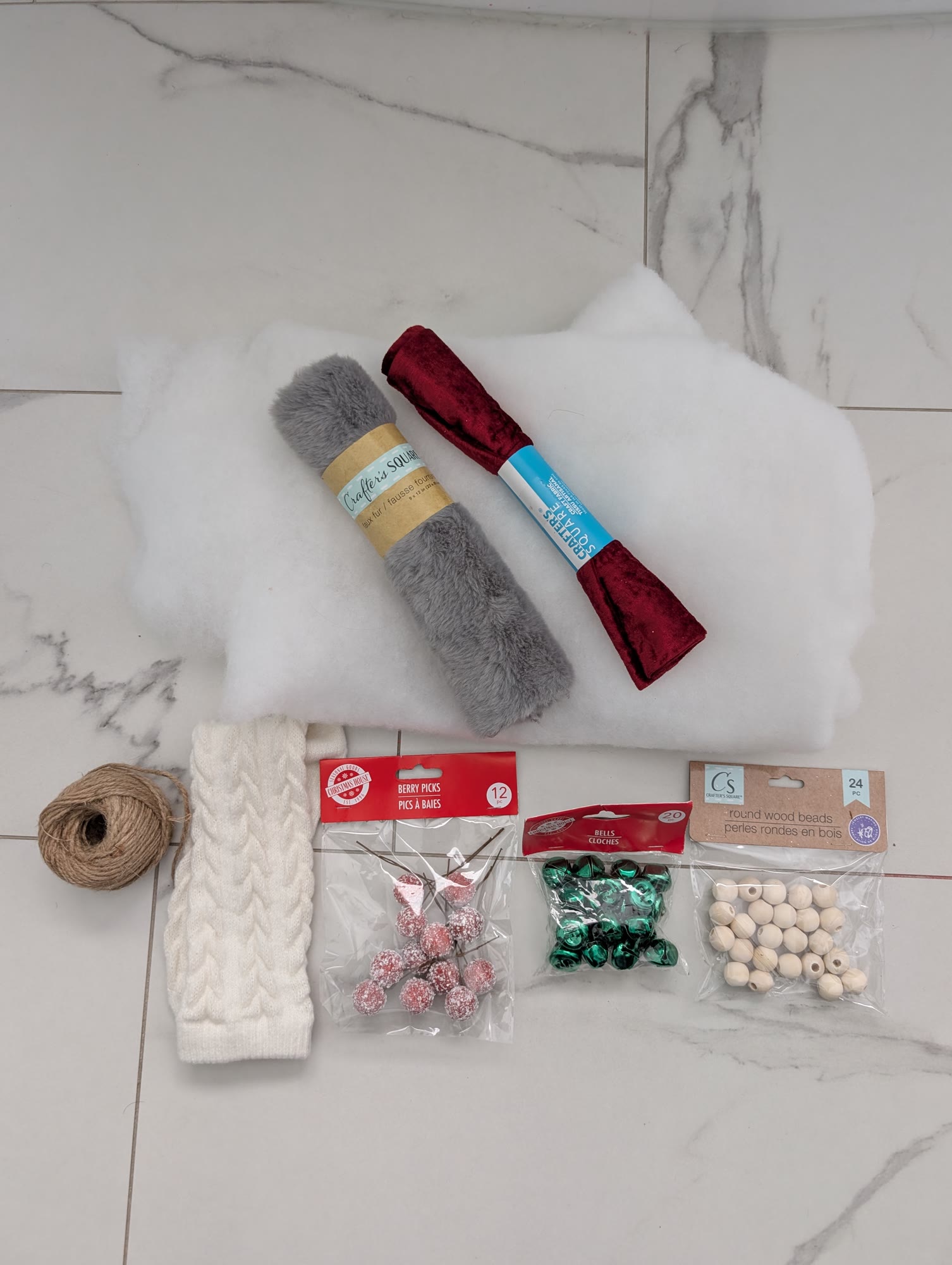Materials on a white tile floor: white stuffing, grey faux fur, red velvet, a cream cable-knit sock, twine, a pack of red berry picks, green bells and wood beads