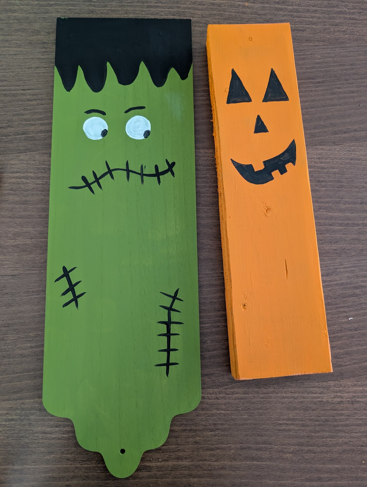 Finished painted green Frankenstein board with black hair, white eyes and stitches, next to an orange pumpkin board with a black jack-o-lantern face
