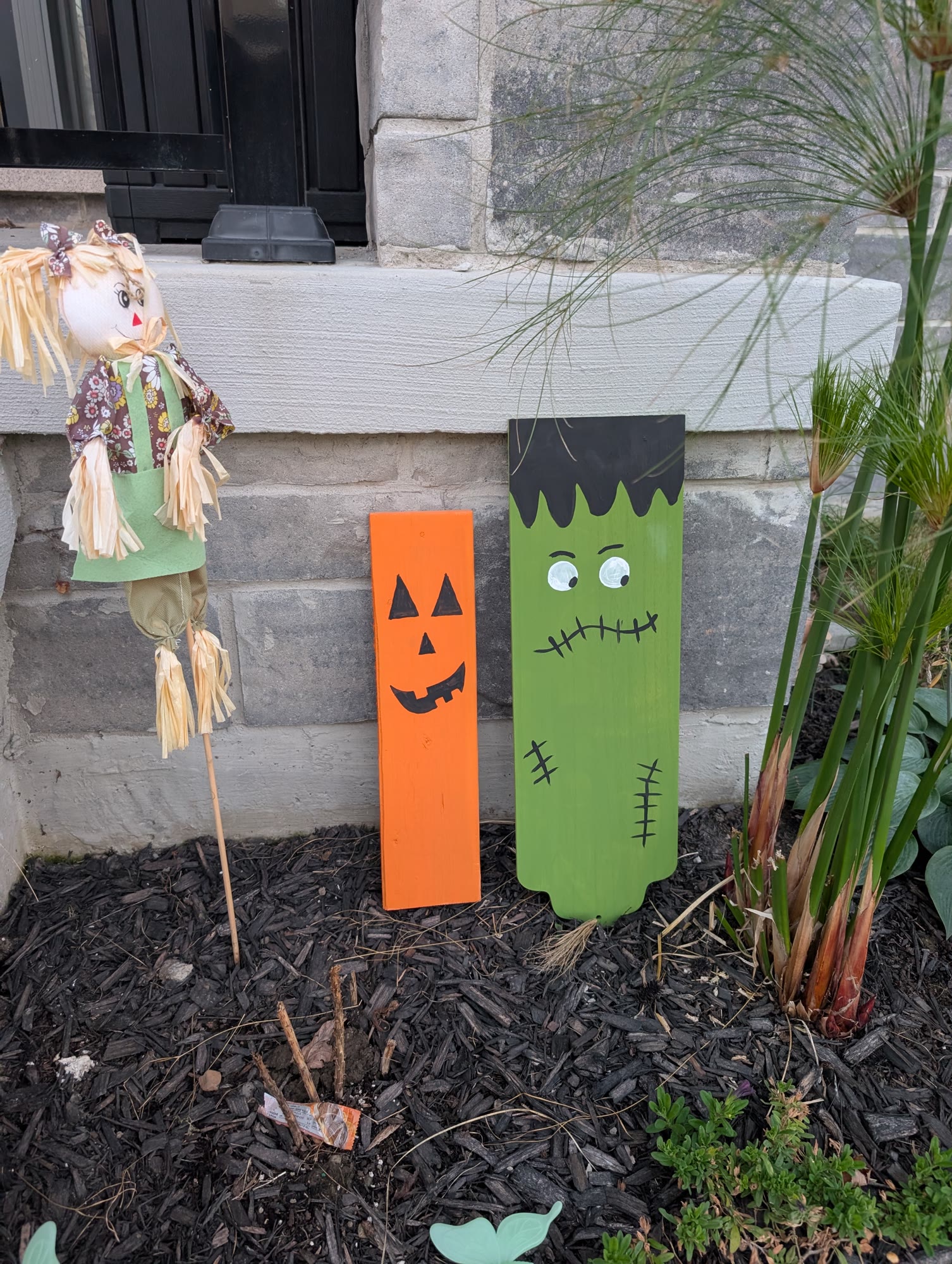 Painted orange jack-o-lantern and green Frankenstein wooden Halloween decorations leaning against a stone wall in a garden bed with a scarecrow