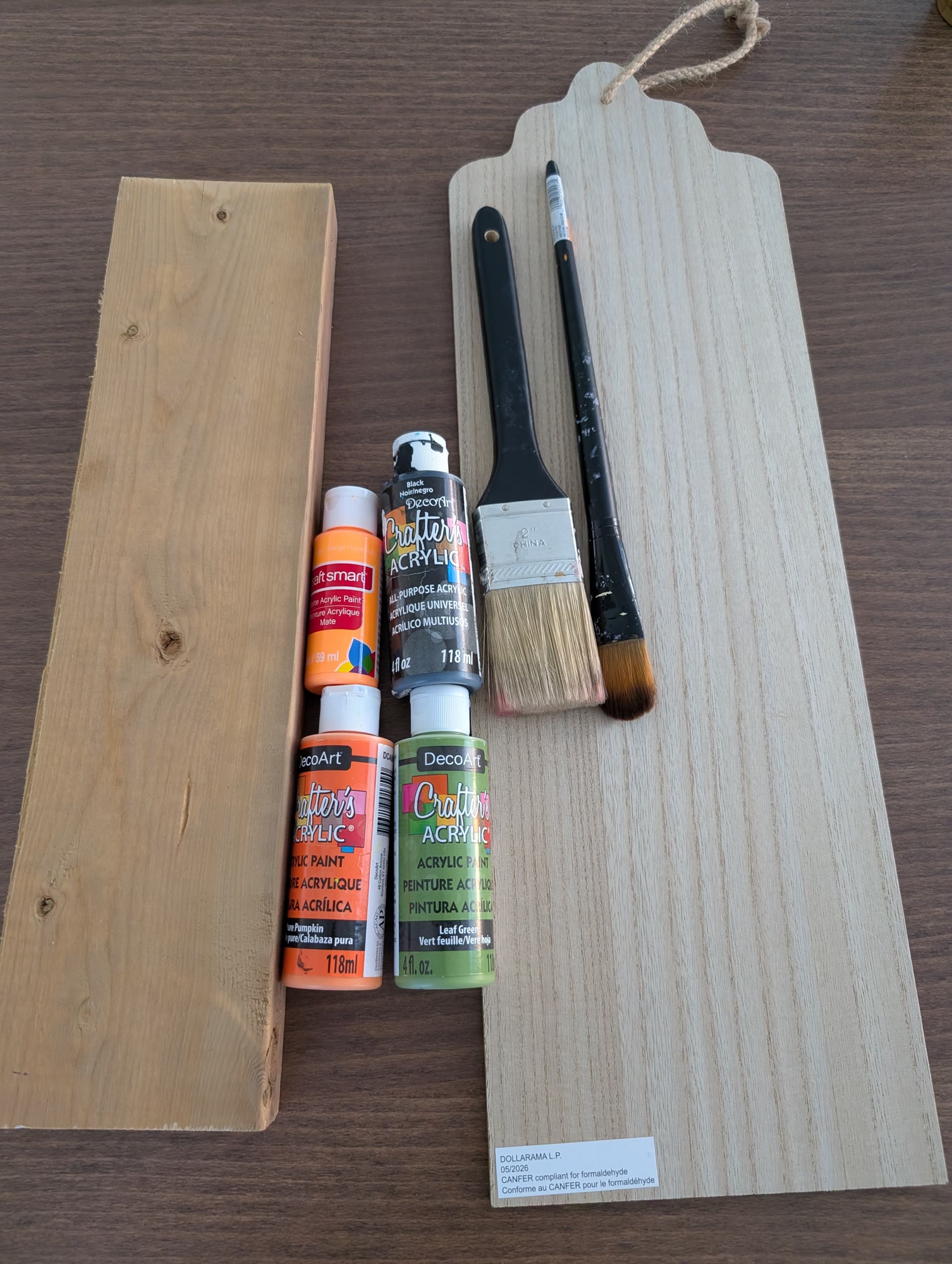 A piece of 2x4 and a Dollarama wood board with bottles of orange, green and black acrylic paint, a wide paintbrush and a small paintbrush