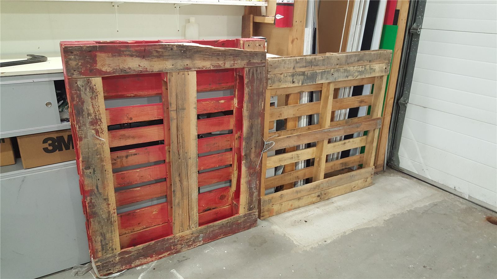 Two disassembled wood skids leaning against a garage wall, one with red paint