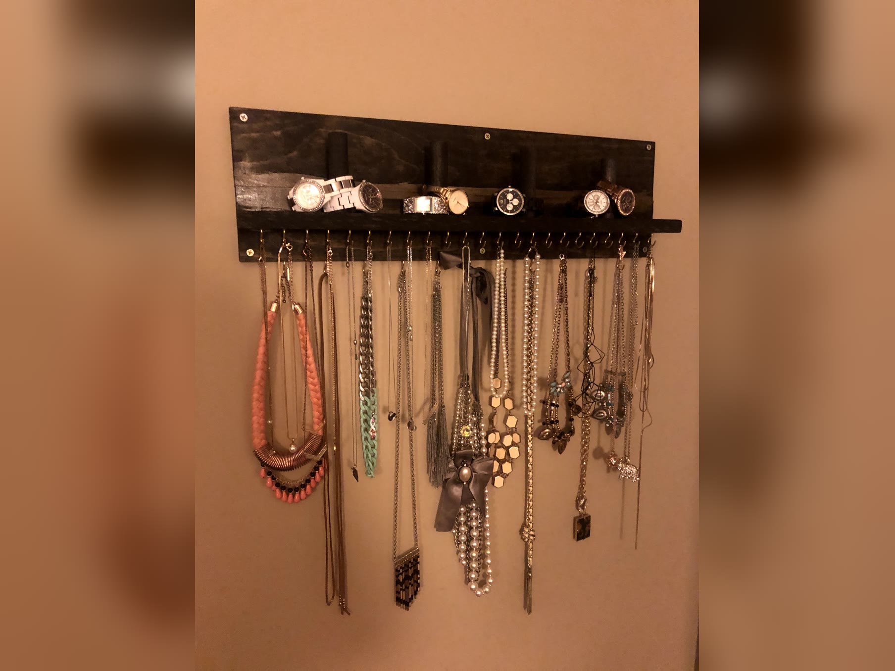 Dark stained wall-mounted DIY jewelry holder with brass hooks holding necklaces and a ledge with dowels holding watches