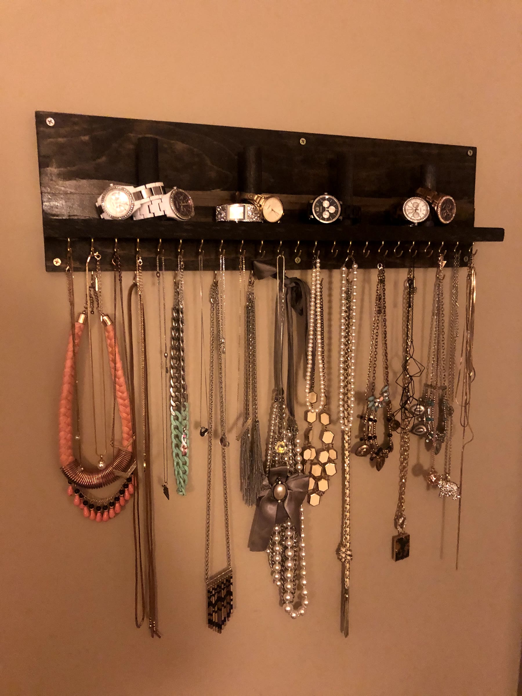 Finished dark stained wall-mounted jewelry holder with a row of brass hooks holding many necklaces and a shelf with four dowels holding watches and bracelets