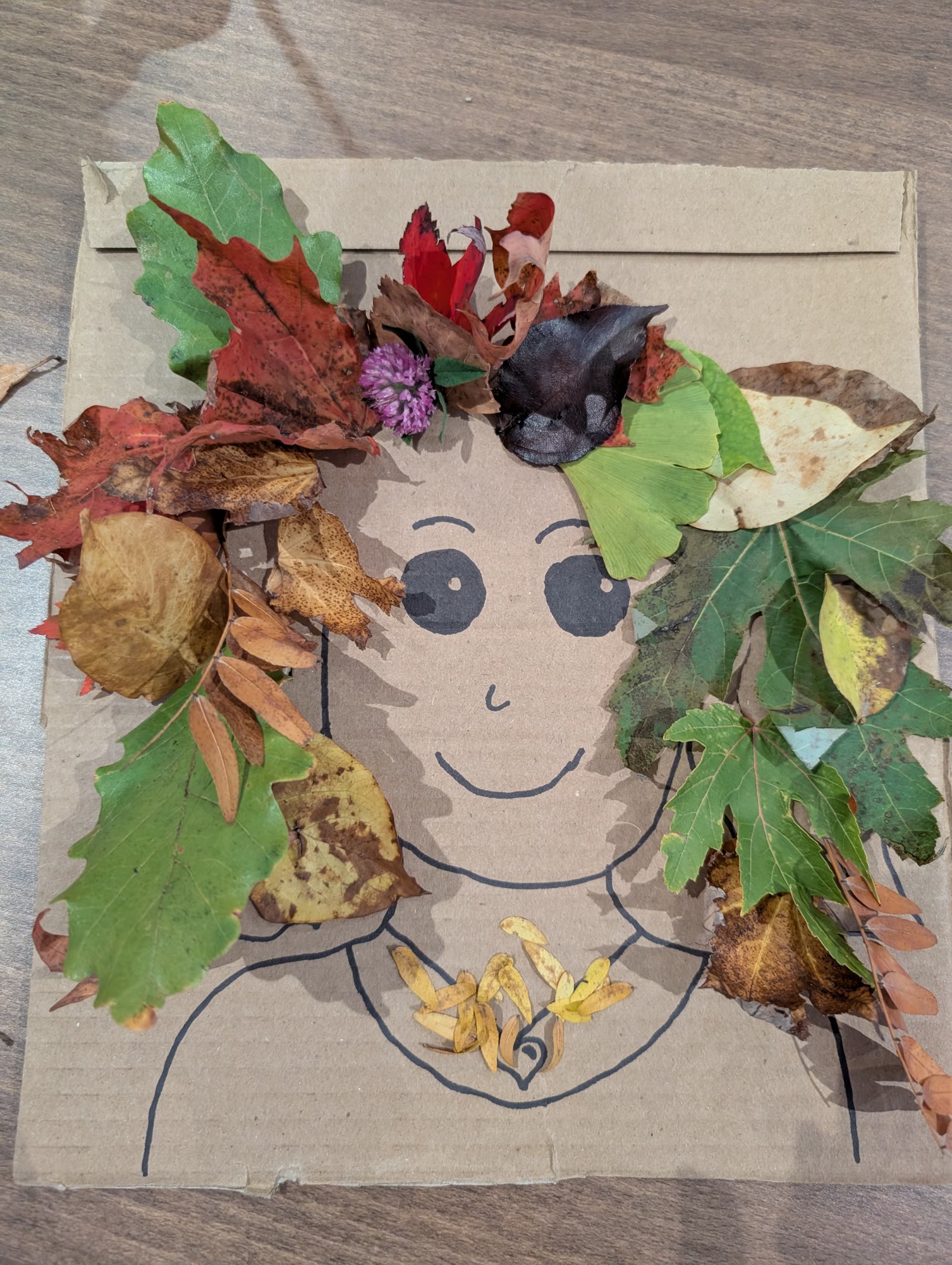 Finished cardboard face with long hair made from green, red and yellow leaves, a clover flower and seed pods
