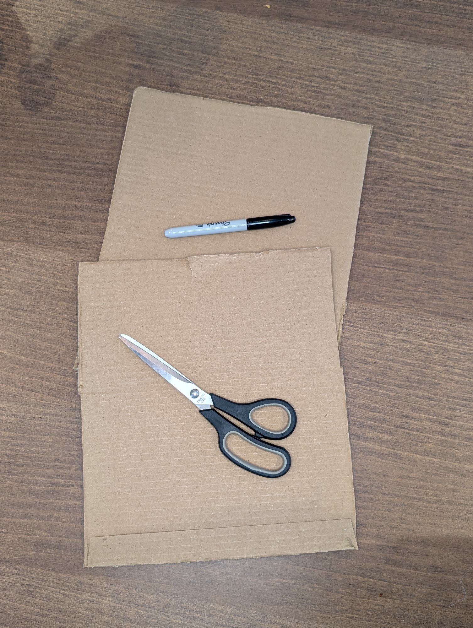 Two pieces of cardboard on a wooden table with a black marker and a pair of scissors on top