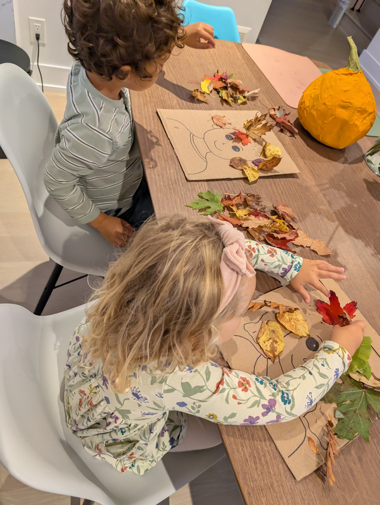 Two kids at a table building leaf hair on cardboard faces, with piles of fall leaves in front of them and a pumpkin in the background