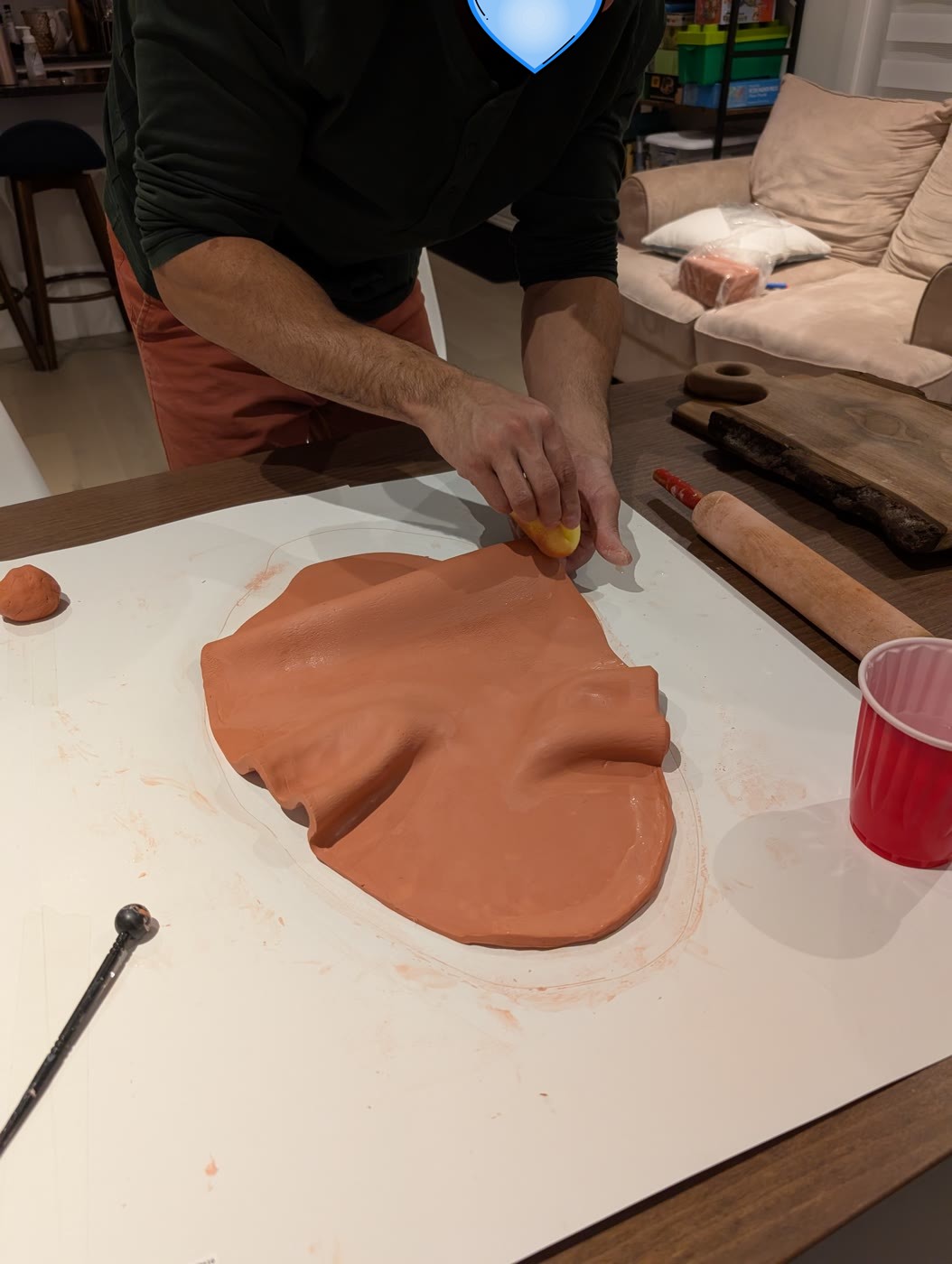 Someone smoothing a large sheet of terracotta clay on a table during DIY ceramic night, with a rolling pin and a cup of water nearby
