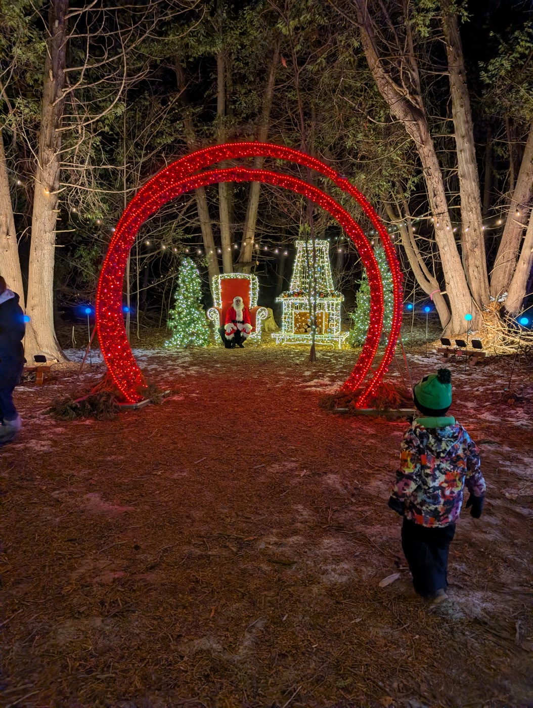 A child in a green hat walking toward a glowing red arch of lights at a festival of lights, with Santa seated in the distance