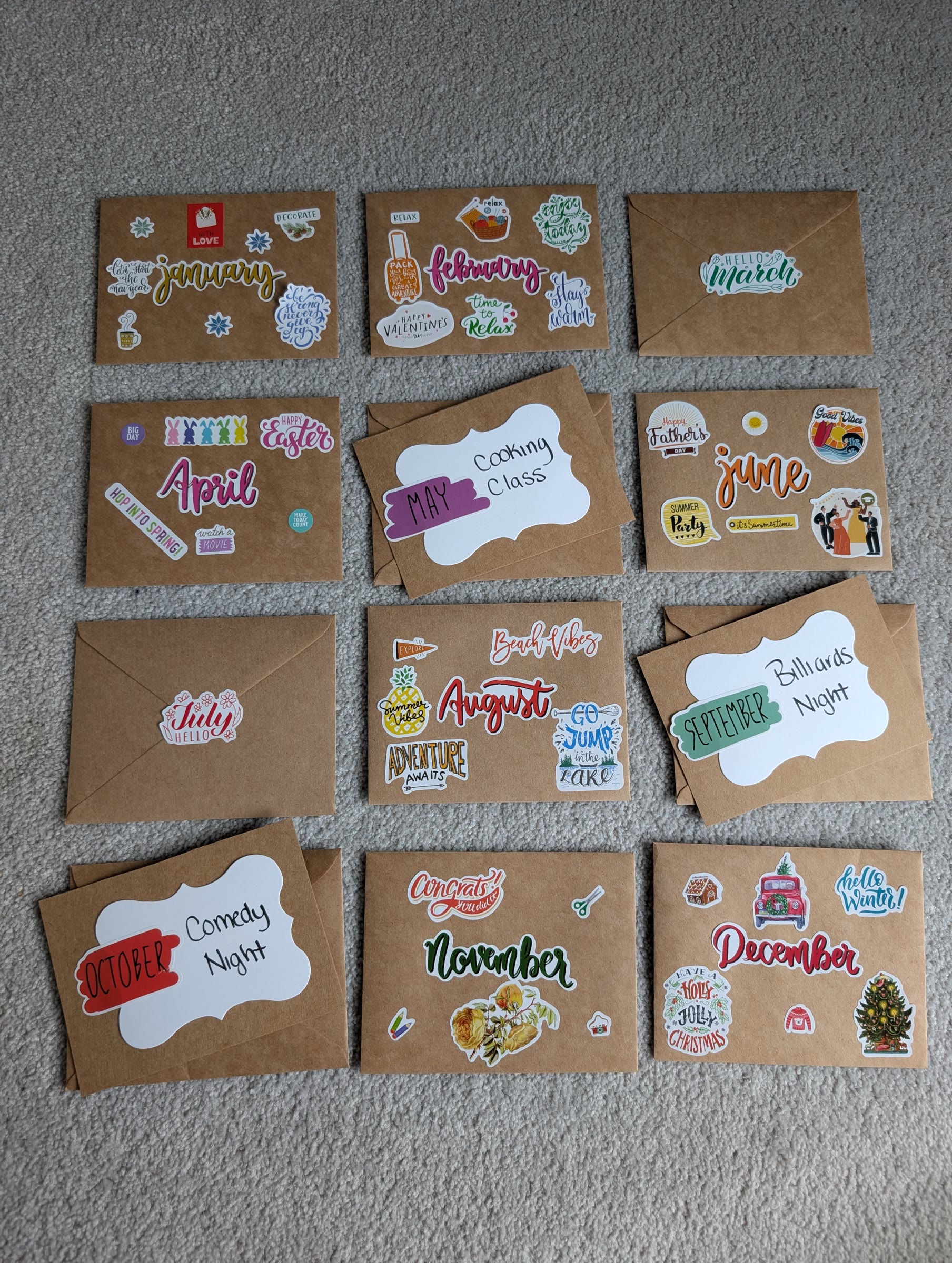 Twelve envelopes laid out in a grid: some with clue stickers on the front, some turned over to show a month sticker sealing the back, and a few with the finished white tag cards on top