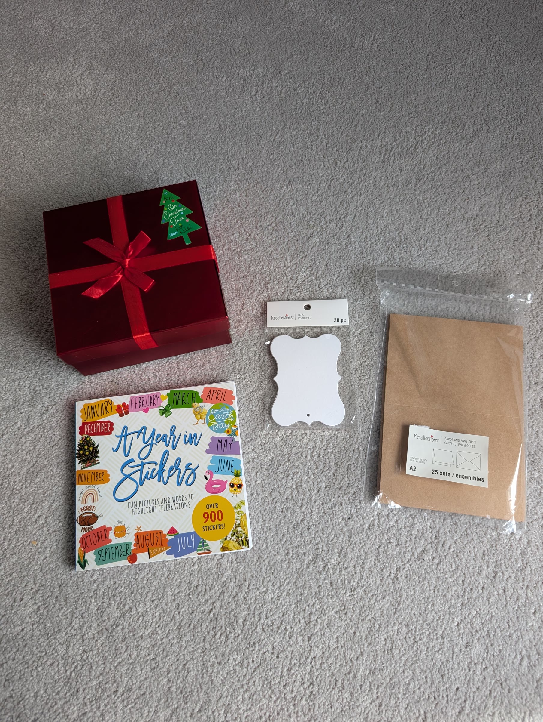 A red gift box, a book of month and holiday stickers called A Year in Stickers, a pack of white ornate gift tags, and a pack of kraft cards and envelopes laid out on carpet