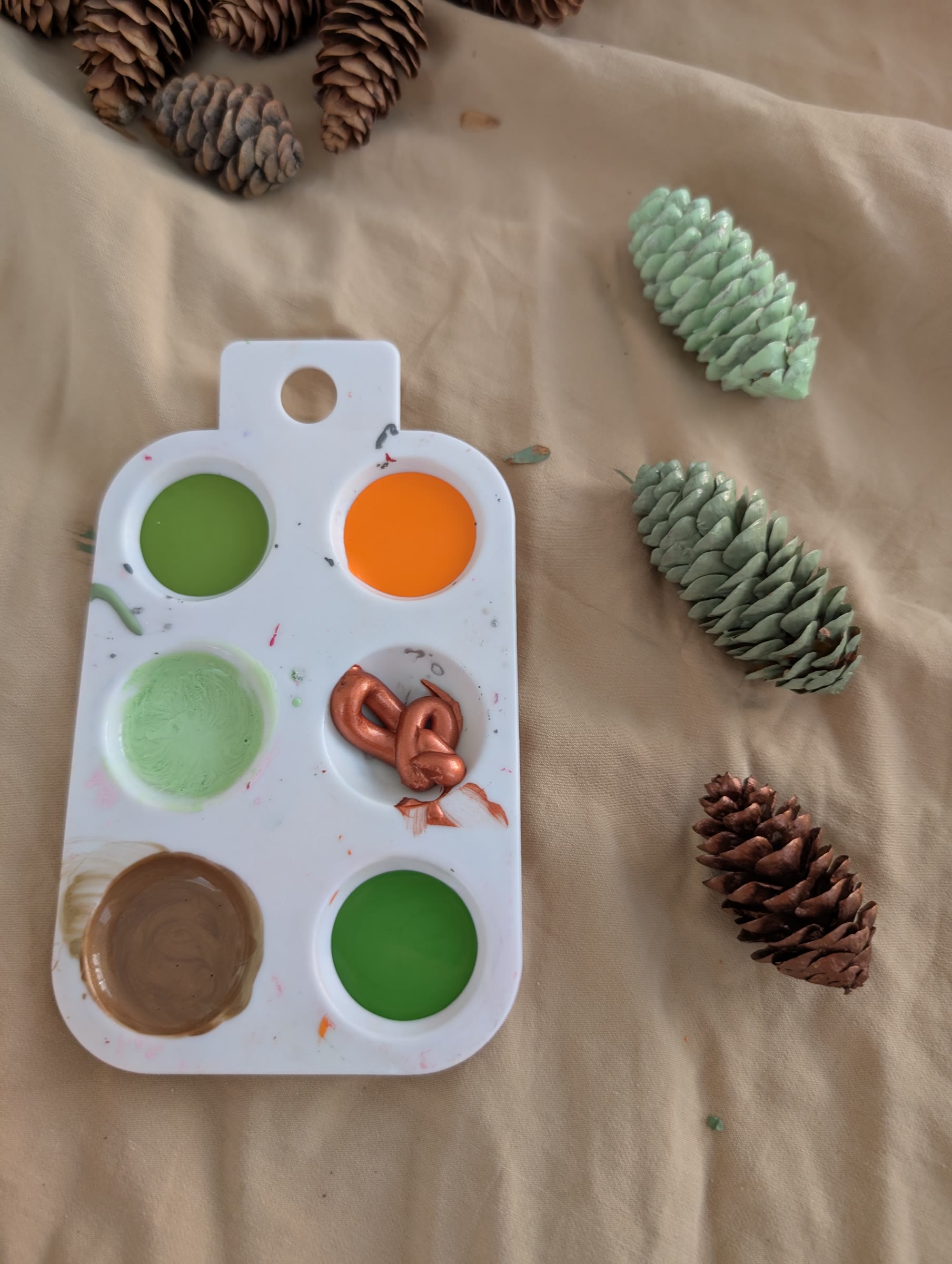 A paint palette with orange, copper, gold, and green paint next to pinecones painted mint green, sage green, and copper