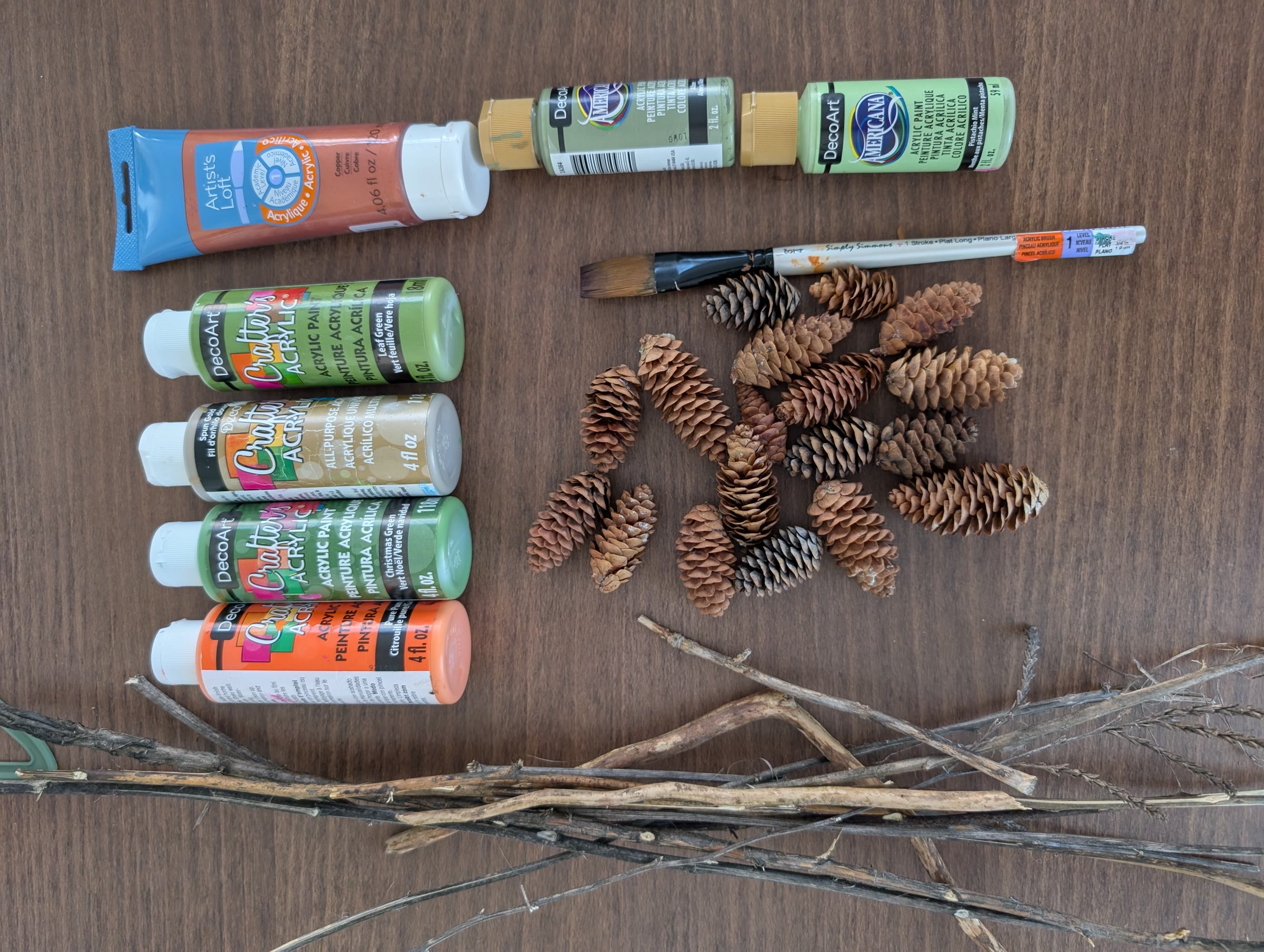 Acrylic paints in oranges, coppers, and greens, a paintbrush, a pile of pinecones, and a bundle of sticks laid out on a wood table