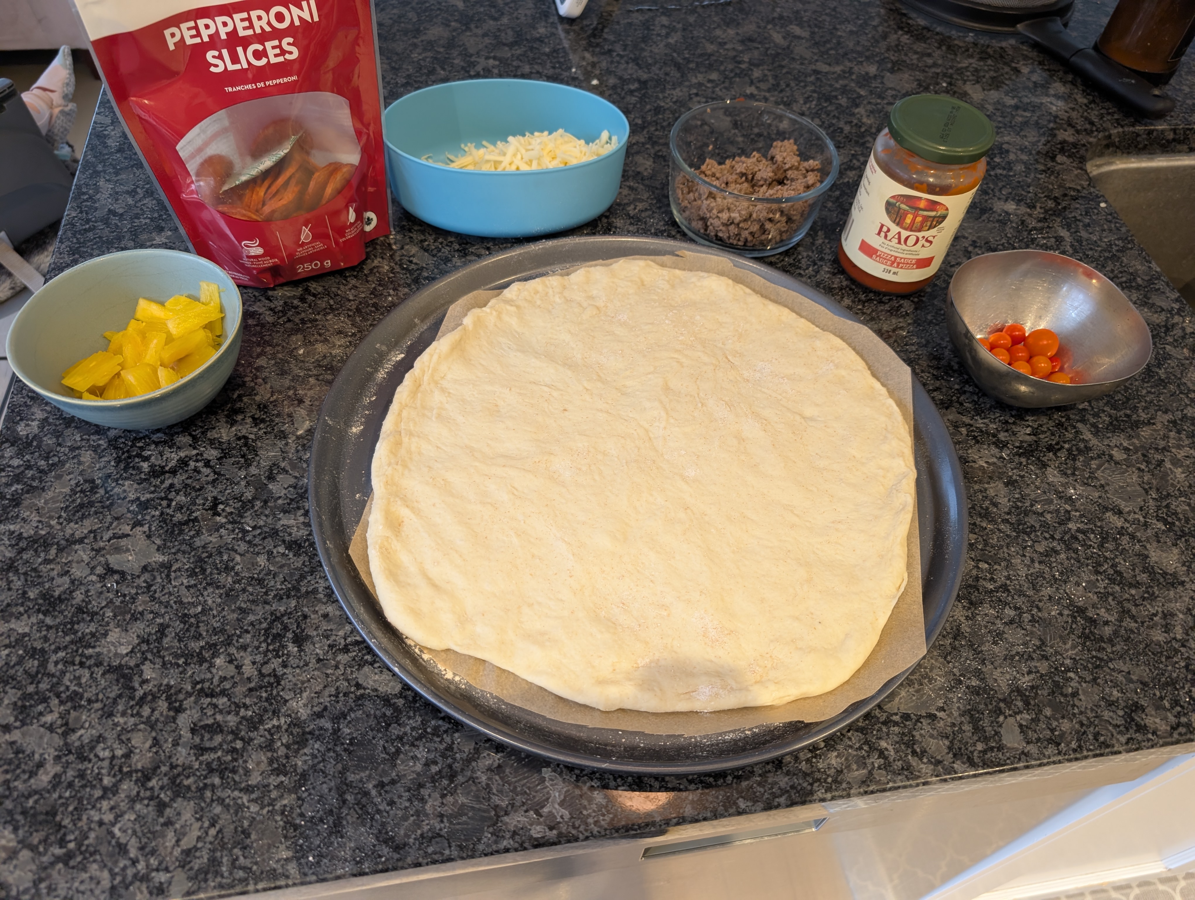 Pizza dough on a pan surrounded by bowls of pepperoni, cheese, ground beef, and pineapple