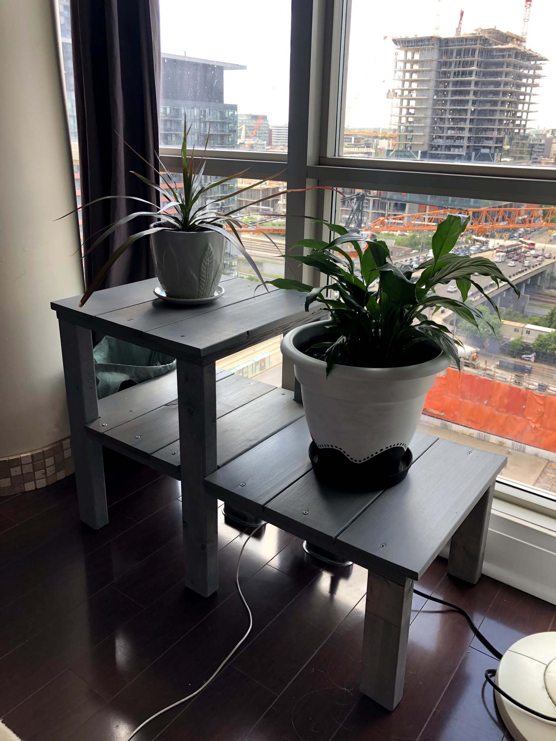 Finished weathered grey two-level plant stand in front of a window, holding a large white potted peace lily on the low level and a spiky plant in a white pot on the tall level