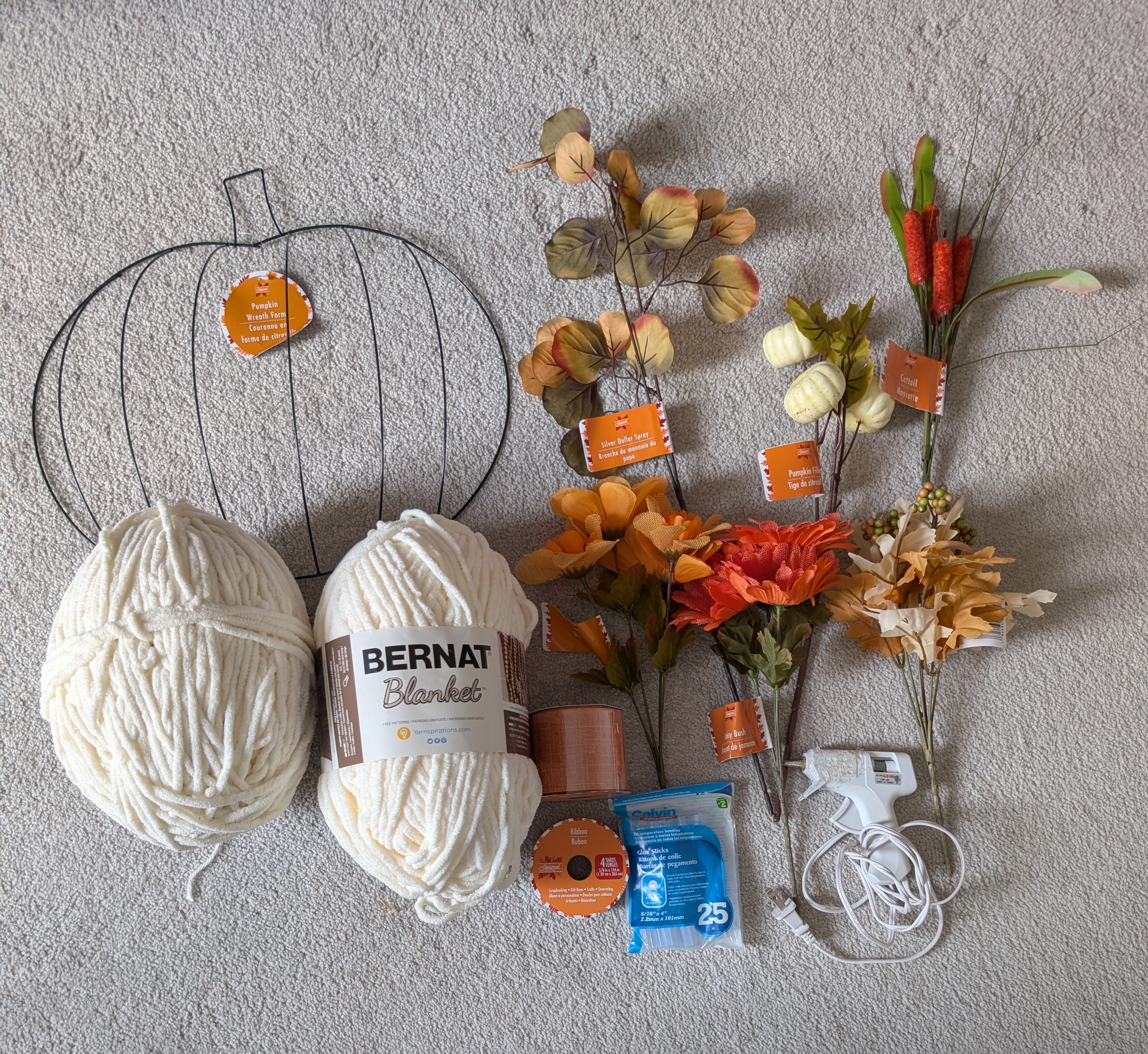 Wire pumpkin frame, cream yarn, fall florals, ribbon, and a glue gun laid out on the carpet