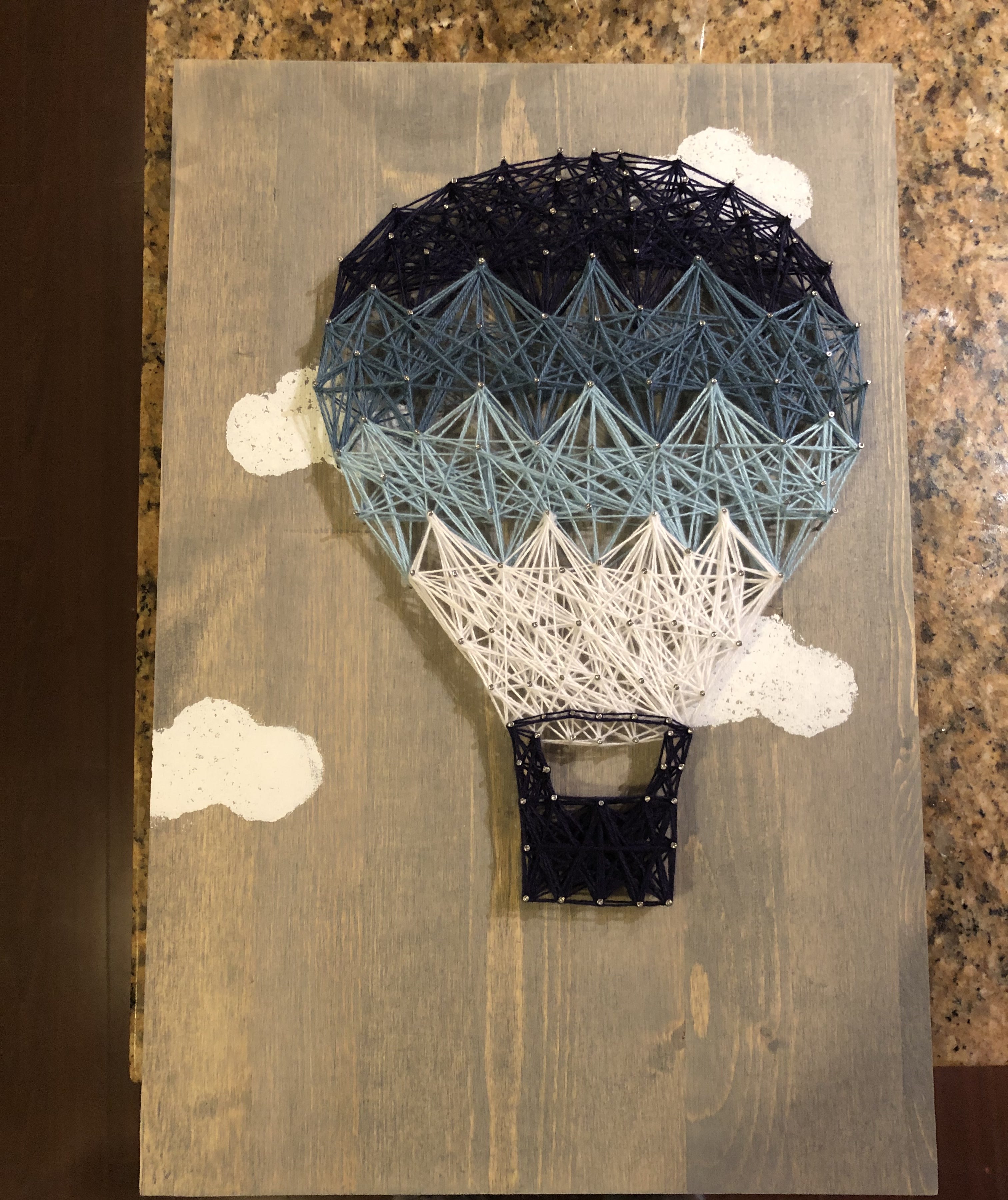 Hot air balloon string art in navy, blue, and white thread on a gray stained board with painted clouds