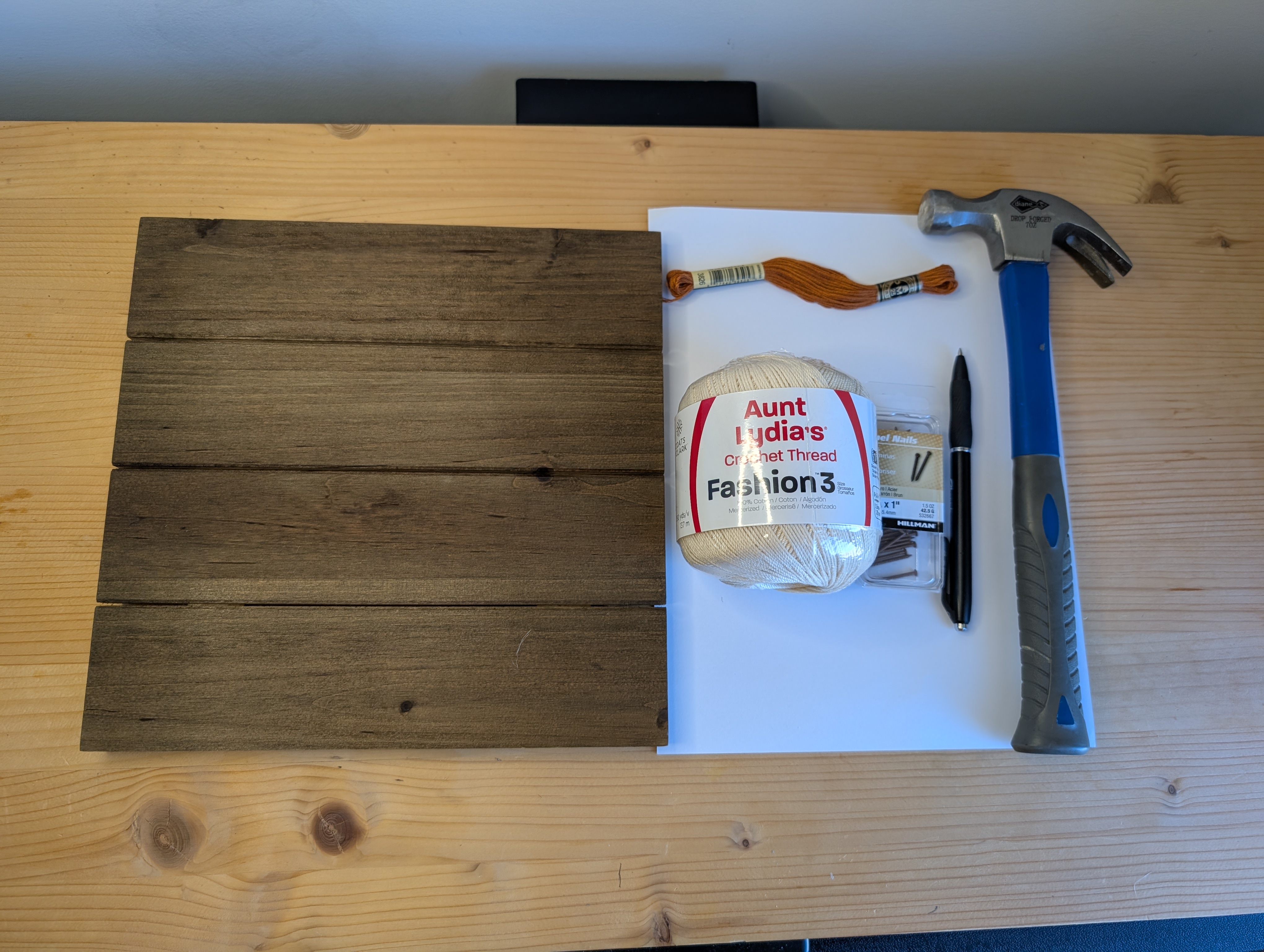 Stained wood plank board, crochet thread, embroidery floss, nails, a hammer, and a pen laid out on a workbench