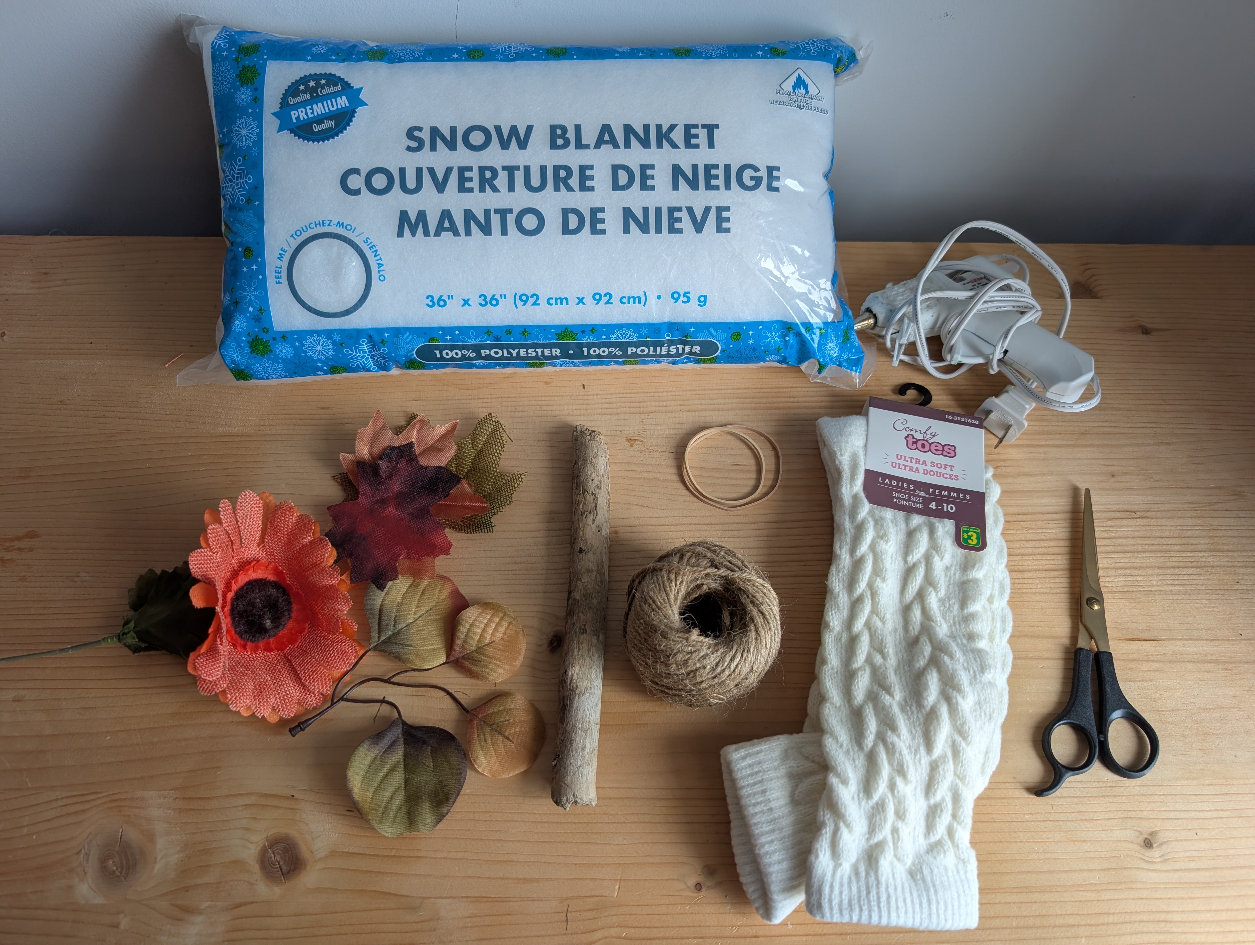 Materials laid out on a wood table: snow blanket stuffing, hot glue gun, faux leaves and flower, a stick, rubber bands, twine, a cream knit sock, and scissors