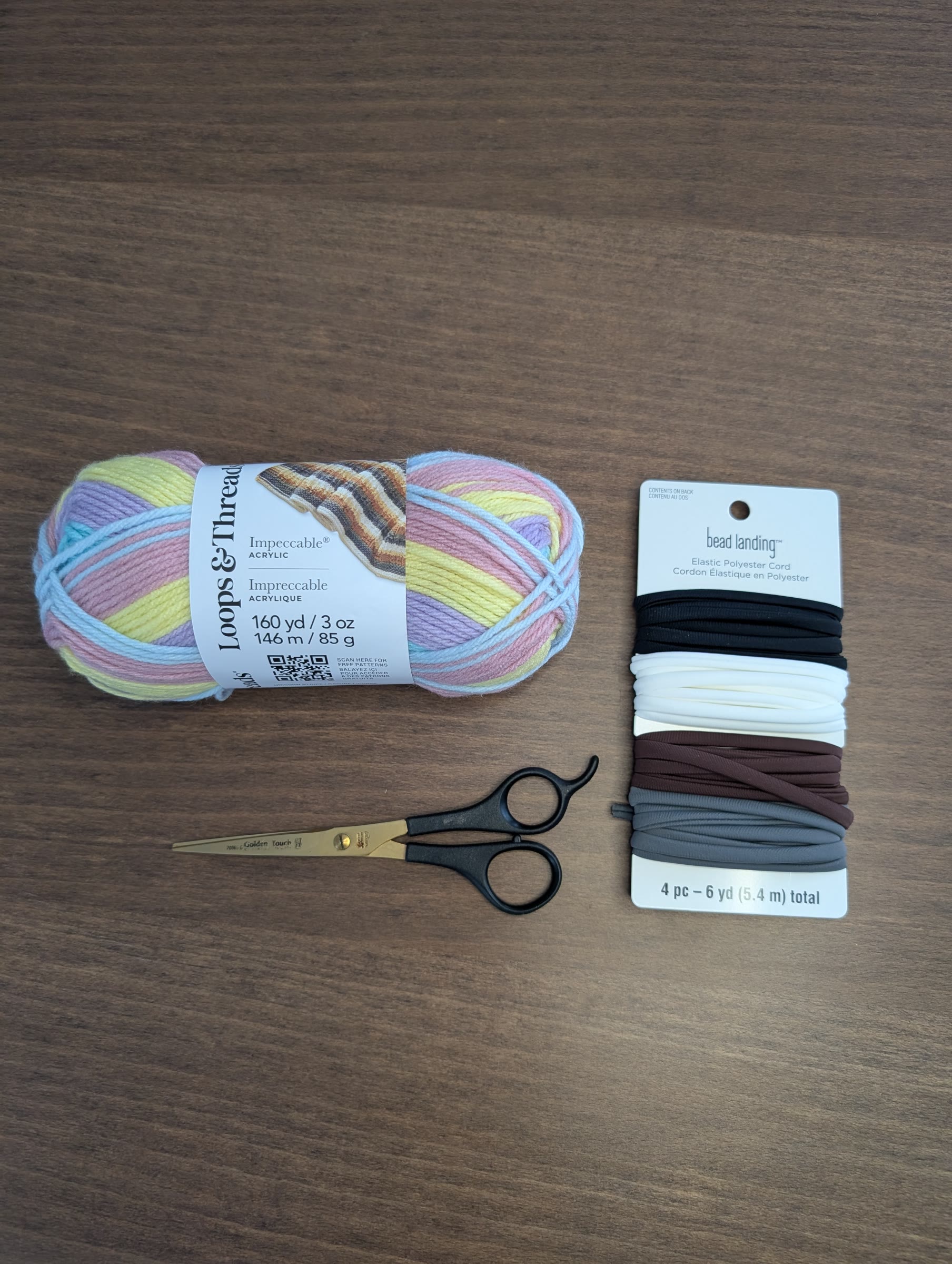 A ball of pastel striped yarn, a card of white, black, brown, and gray elastic cord, and a pair of scissors on a wood table