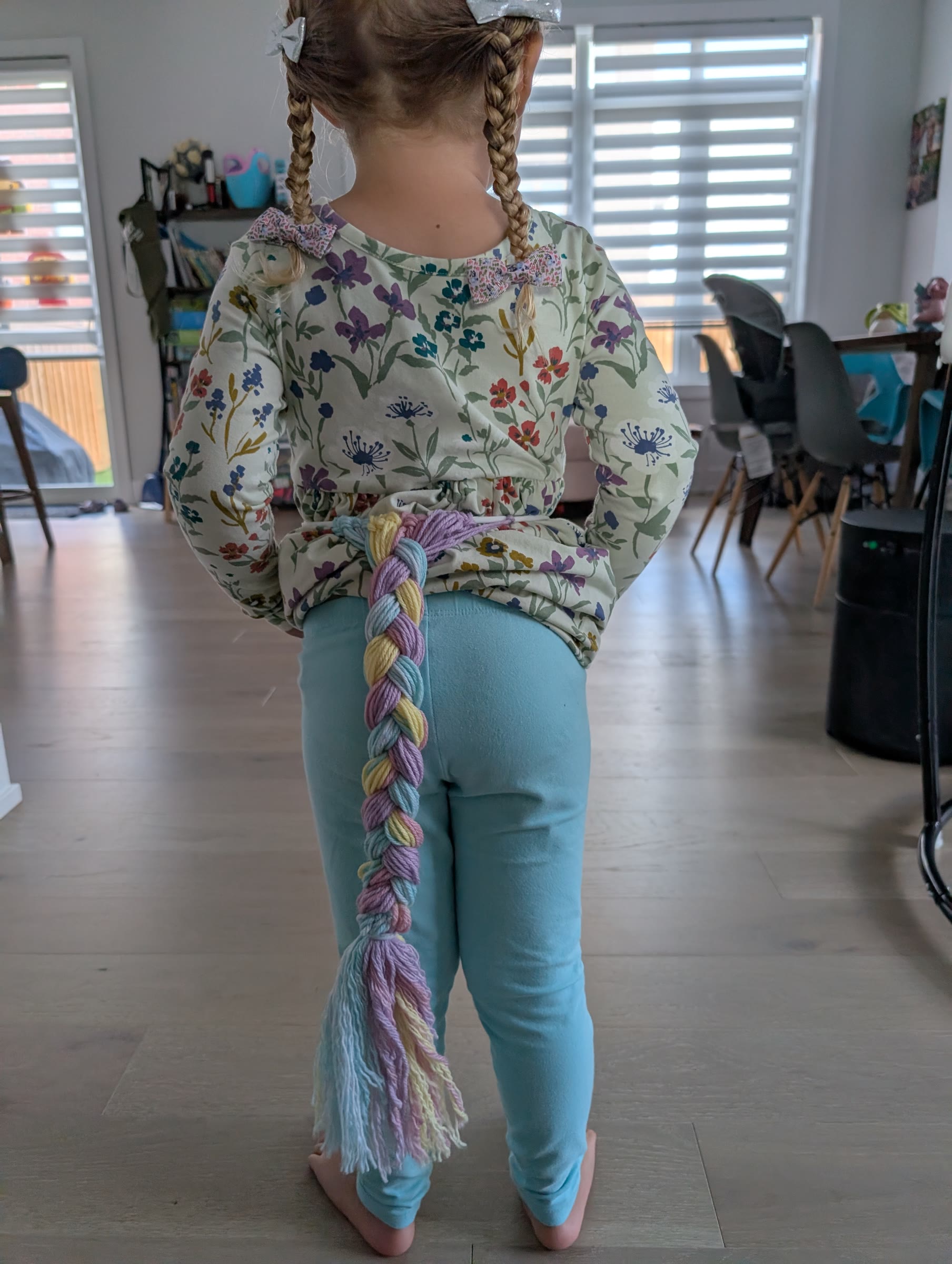 Child from behind wearing a braided pastel rainbow yarn unicorn tail with a fringed end, tied at the waist